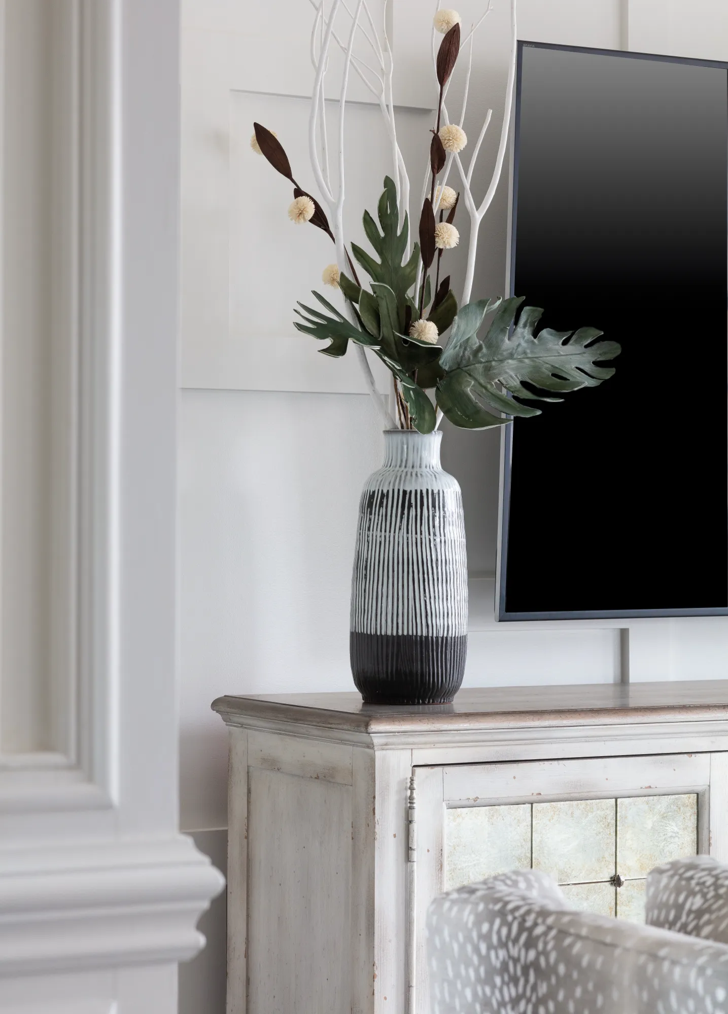 A stylized black and white patterned vase with decorative botanical accents sits on a distressed white media console in a living room, partially obscuring a television.