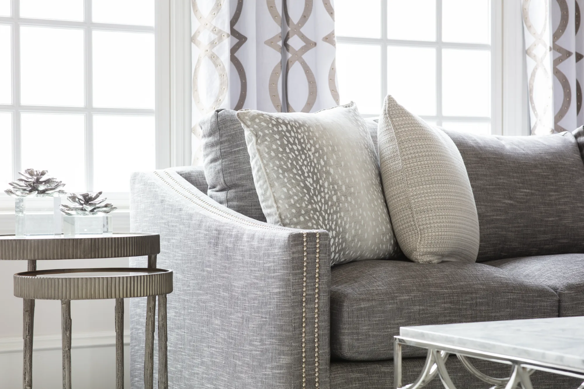 A close-up of an elegant gray sofa with silver nailhead trim and patterned pillows. A tiered silver side table with decor is beside the bright, gridded window.