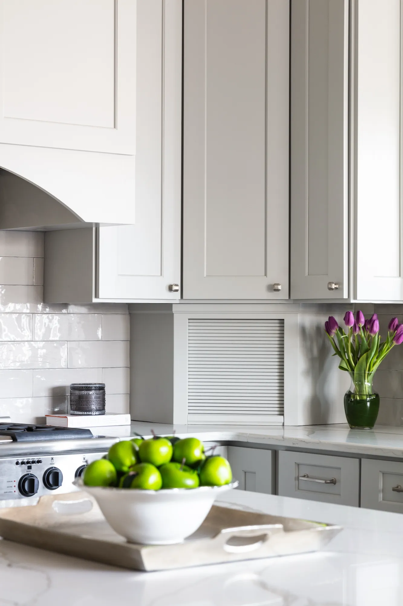An elegant kitchen design featuring gray cabinets, a white tile backsplash, and marble counters. A bowl of green fruit and purple tulips sit prominently on the counter.
