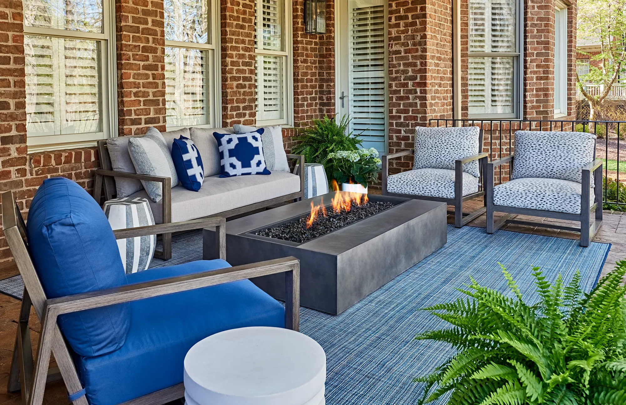 A cozy, modern outdoor patio furnished with blue and gray seating, a woven blue rug, and a rectangular gray gas fire pit burning against a red brick wall.