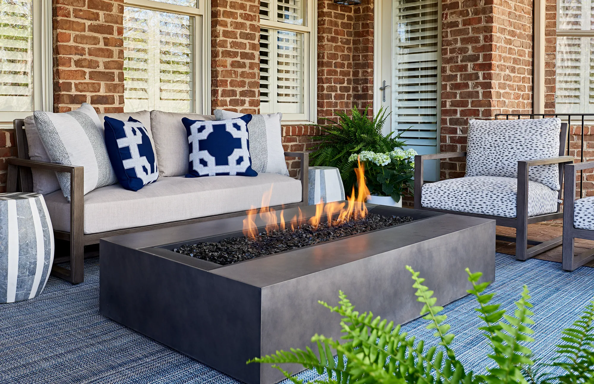 A modern outdoor fire pit burns brightly in the center of a covered brick patio, surrounded by comfortable gray seating and blue patterned pillows.
