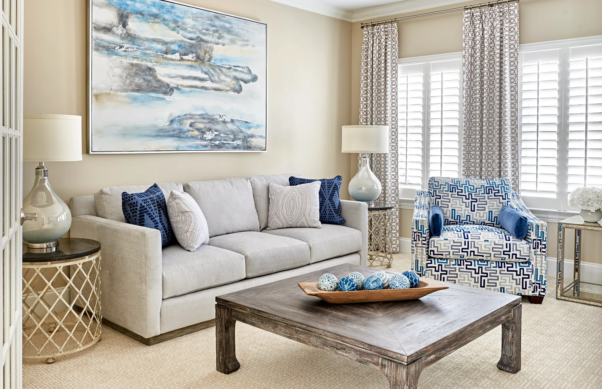 A comfortable living room setup showing a gray sofa with blue pillows, facing a dark wood coffee table. A blue and white geometric patterned armchair sits near a window.