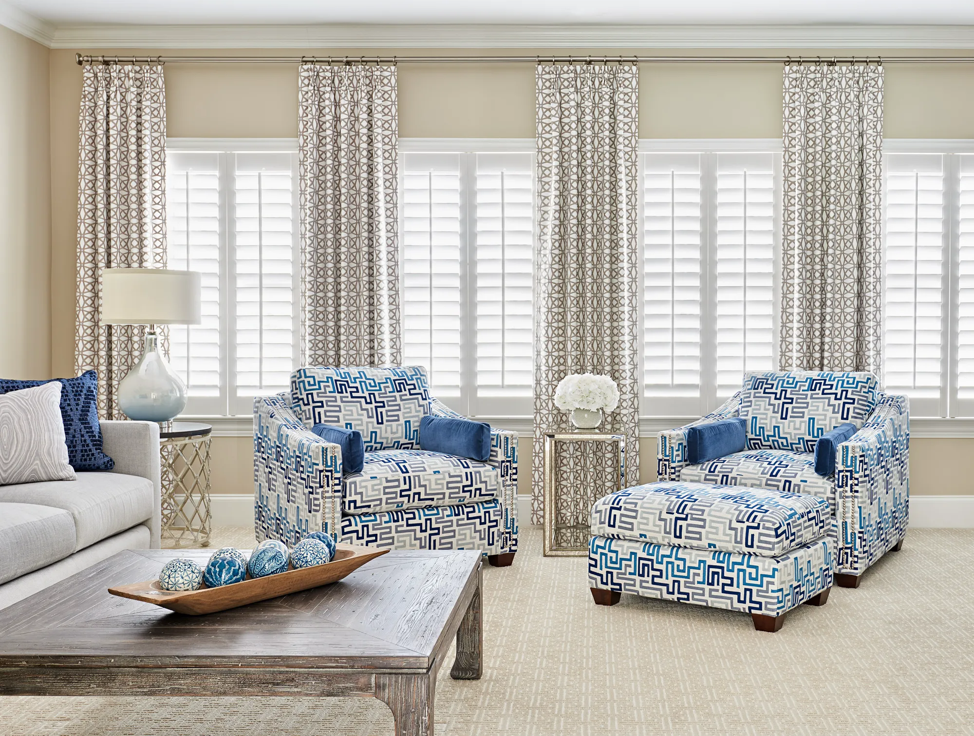 A sophisticated living room featuring two armchairs and an ottoman upholstered in a bold blue geometric pattern. The windows have shutters and patterned drapes.