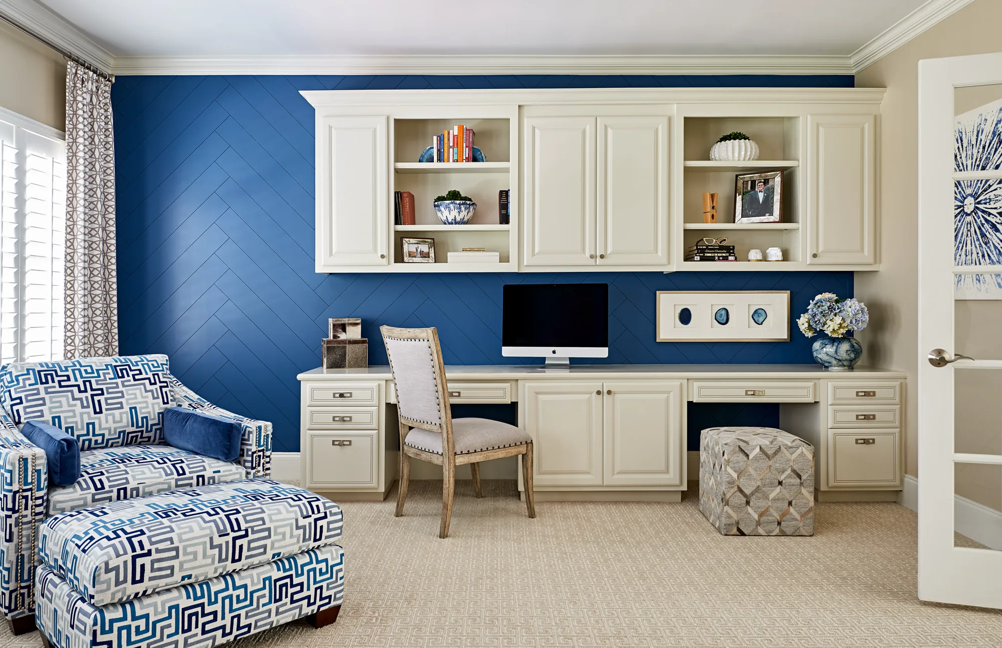 A luxurious blue and white home office featuring built-in white cabinetry against a vibrant blue wall with a chevron pattern. A patterned armchair and ottoman are visible.