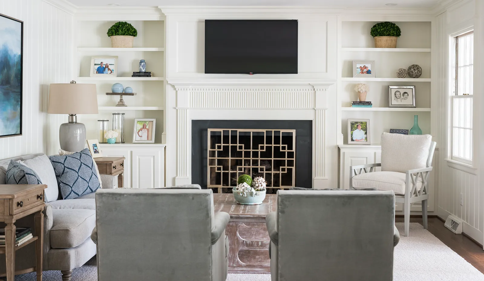 A bright, traditional living room centered on a white fireplace mantel with a TV above it. Built-in shelves flank the fireplace, displaying photos and decor.
