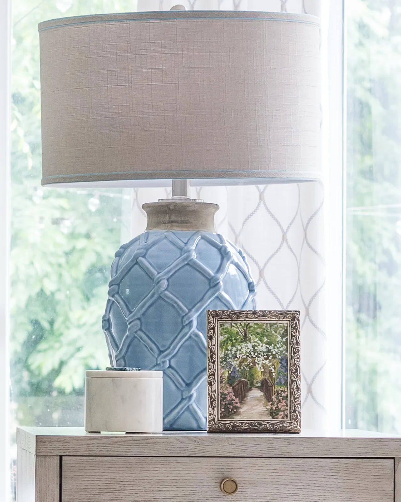 A blue ceramic lamp base with a lattice pattern and a tan drum shade sits on a wooden dresser. A marble container and a small framed painting rest next to the base.