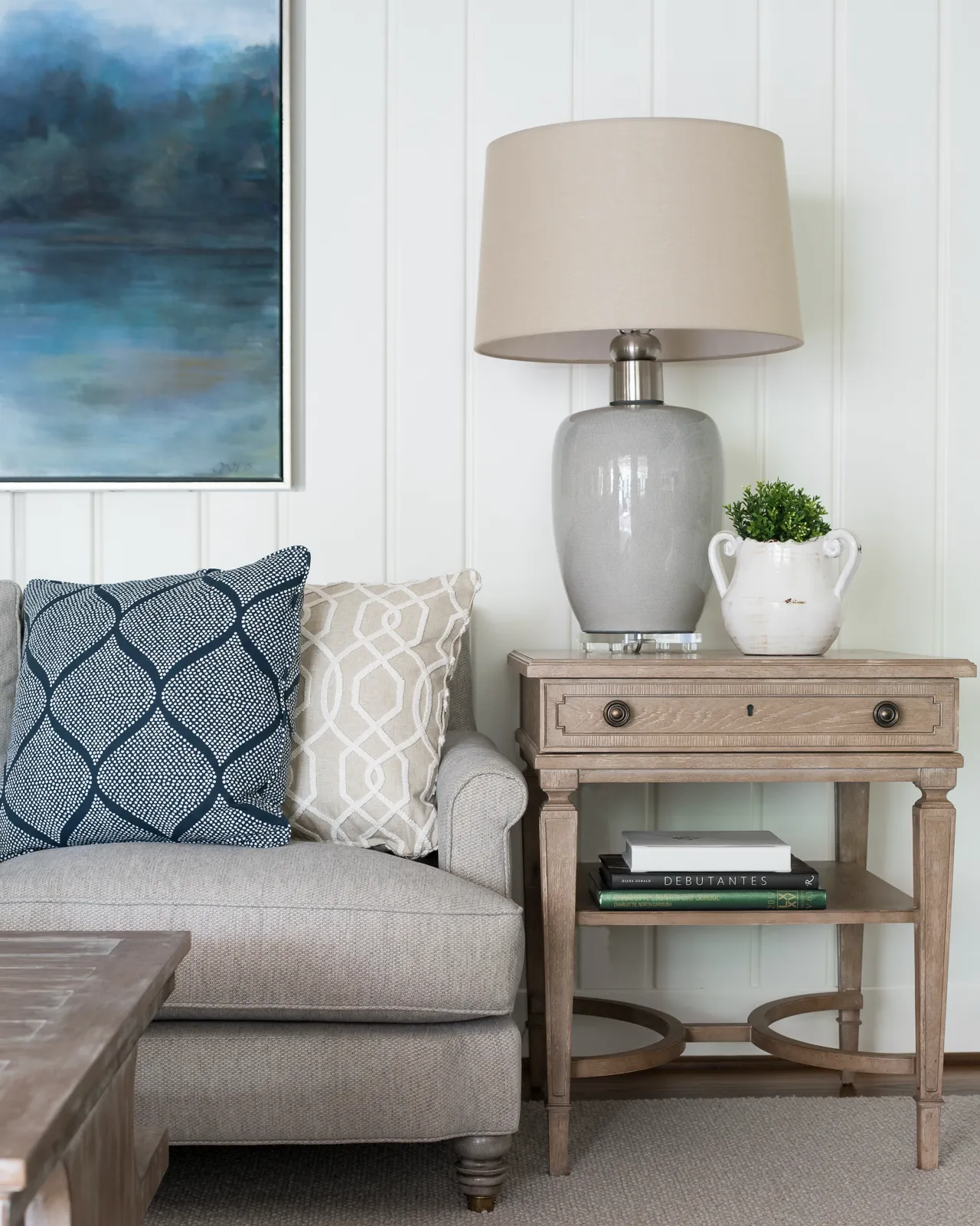 A light gray sofa with two patterned pillows sits next to a wooden side table topped with a gray lamp and a small plant, set against a white paneled wall.
