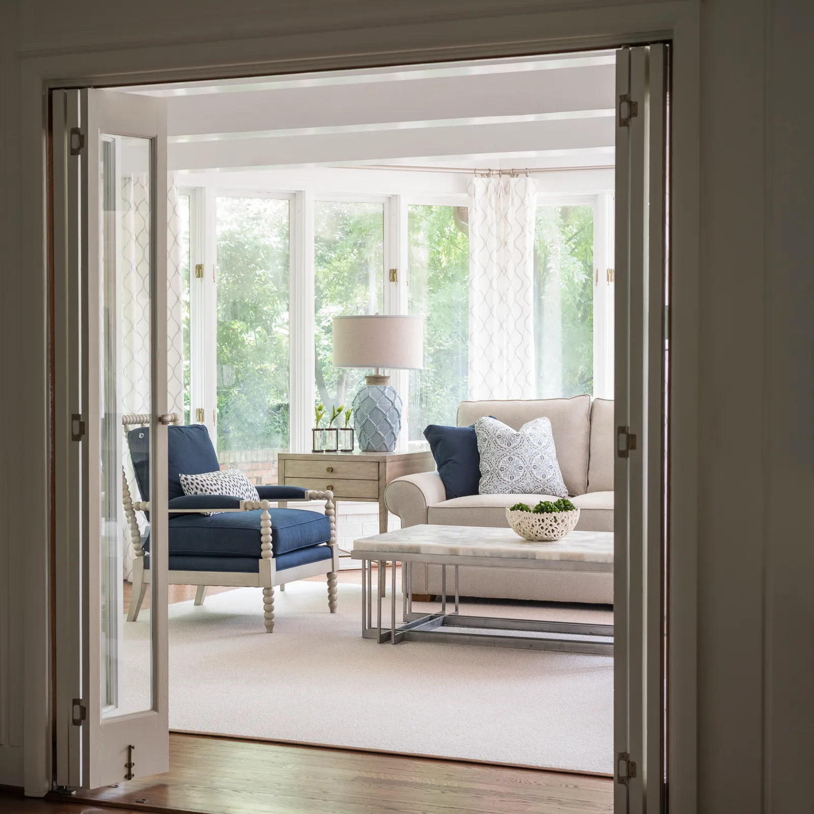 A sunroom with bright windows and lush greenery, seen through open French doors, features a beige sofa and a navy blue armchair on a light rug.