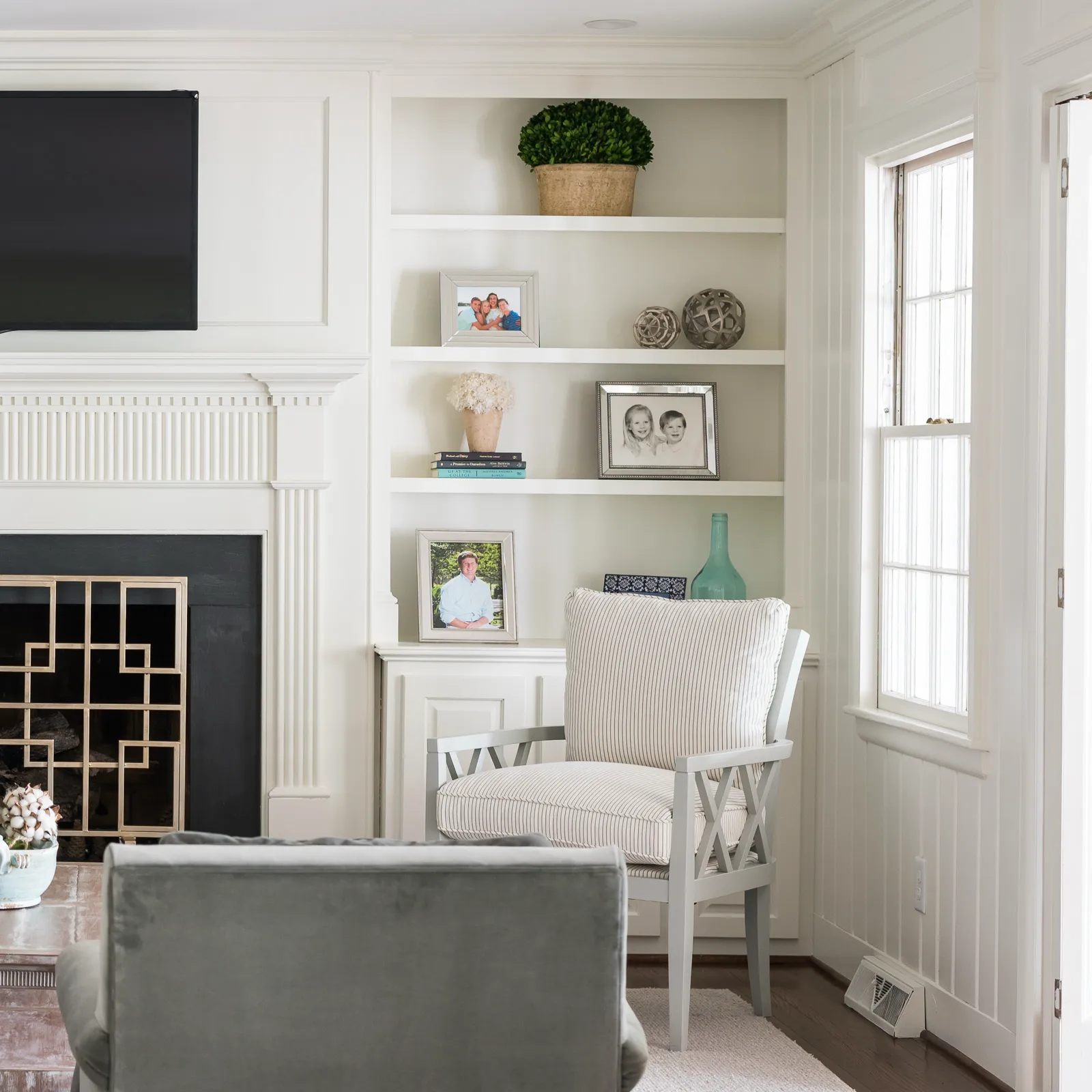 A bright living room corner featuring white built-in shelving with photos, a fireplace, and a gray armchair with striped cushions next to a paneled window.