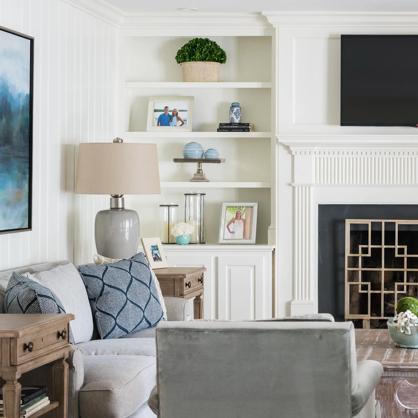 Built-in white shelves and a fireplace anchor this living room. The shelves hold family photos and decor; a grey sofa with patterned blue pillows is in the foreground.