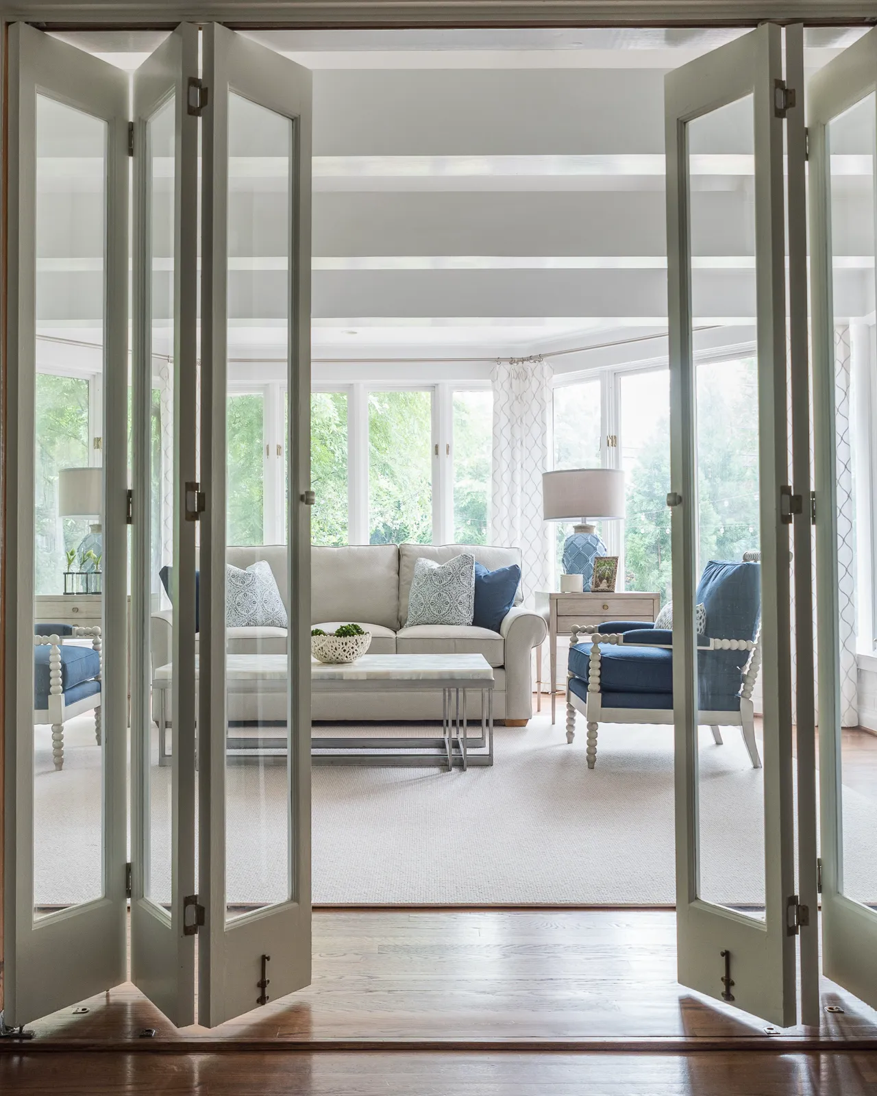 Open folding doors frame a bright living room featuring a neutral sofa, blue accent chairs, and pillows, centered on a modern marble coffee table.