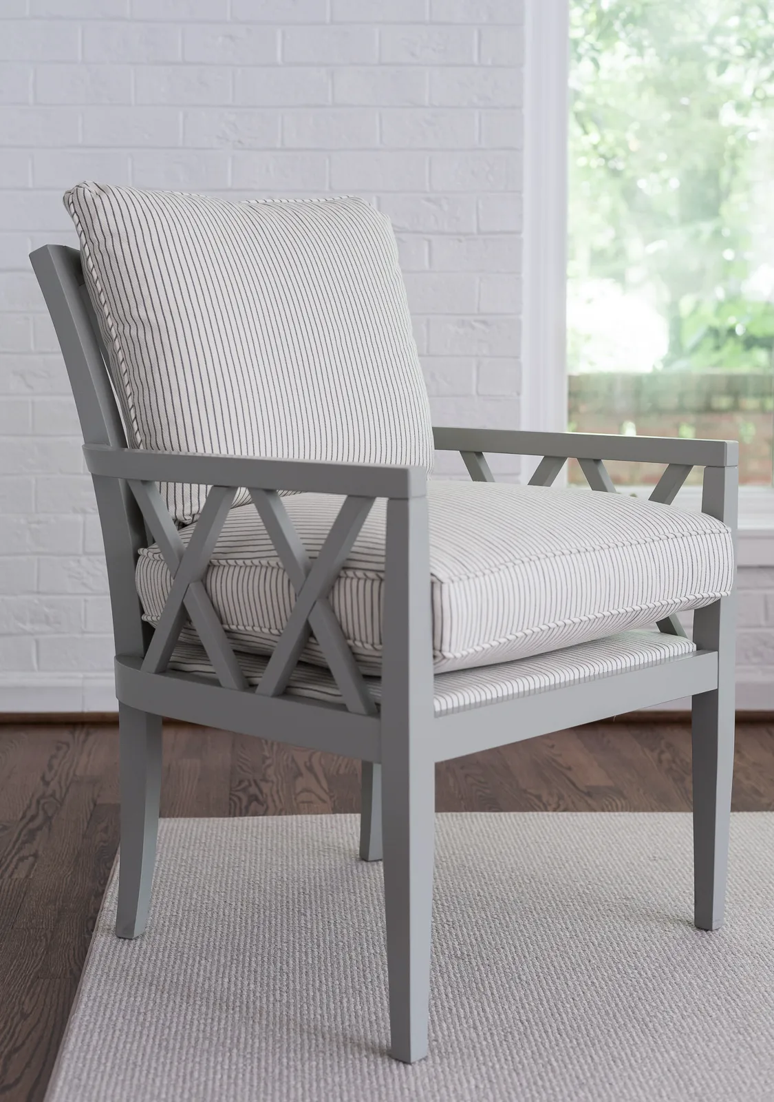 A comfortable light gray patio chair with thick white and black pinstripe cushions rests on a neutral rug in a room with a whitewashed brick wall.