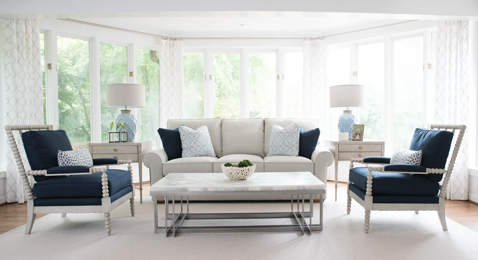 A light and airy living room with a cream sofa and two matching blue armchairs facing a marble coffee table in front of bright bay windows.
