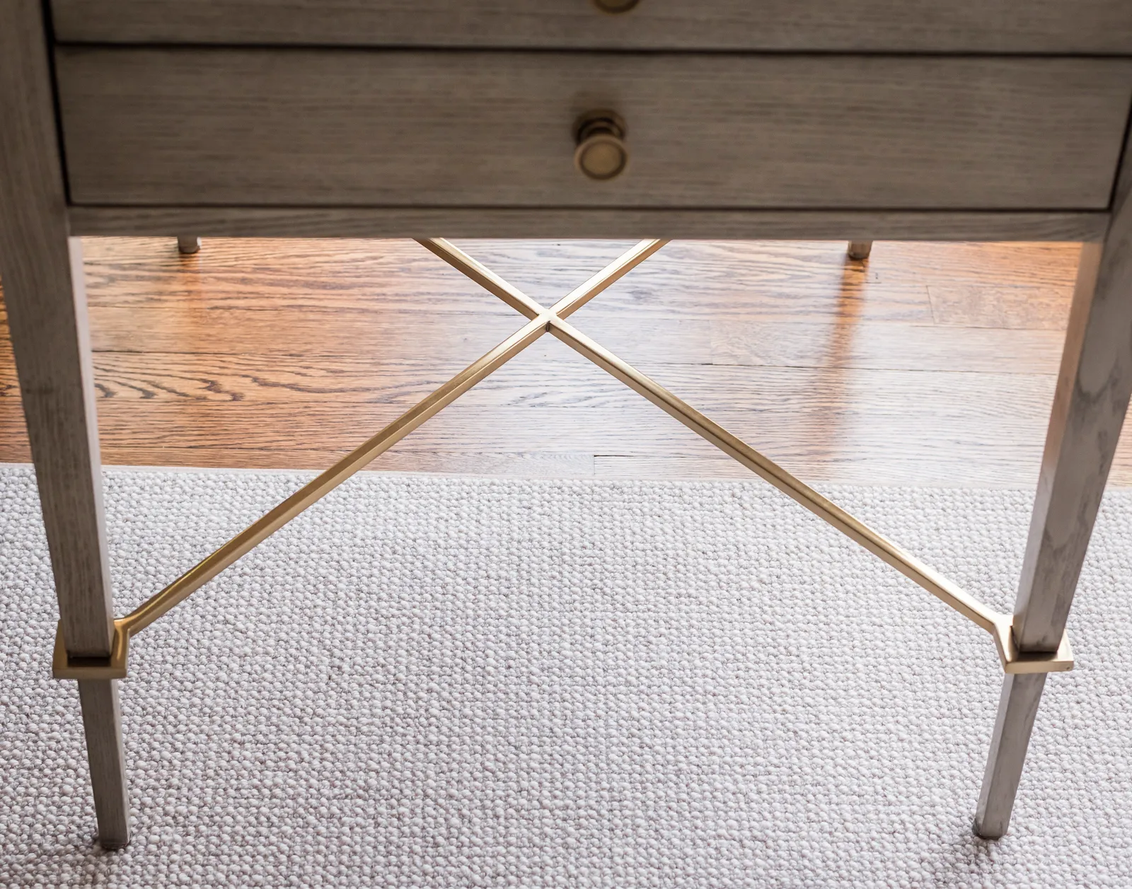 A detailed view of a table's base showing light wood legs, brass metal accents, and an X-shaped stretcher bar. The furniture rests on a light woven area rug.