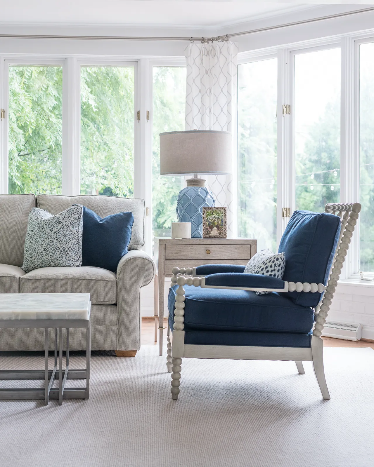 A light-filled sitting area with a textured beige sofa and a striking blue armchair featuring a white bobbin frame. A light blue ceramic lamp sits on a wooden side table near large windows overlooking trees.