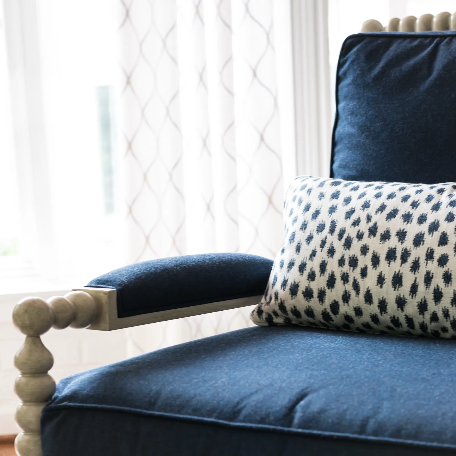 A detail shot of a sophisticated dark blue upholstered armchair. It has a light wooden frame with a spindle design and is accented by a spotted blue and cream pillow.