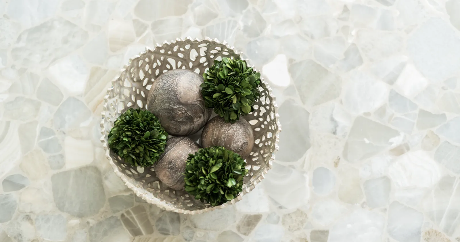 A silver open-work bowl contains three metallic balls and three small green ornamental spheres. The bowl sits on a white countertop made of large, irregular pieces of stone.