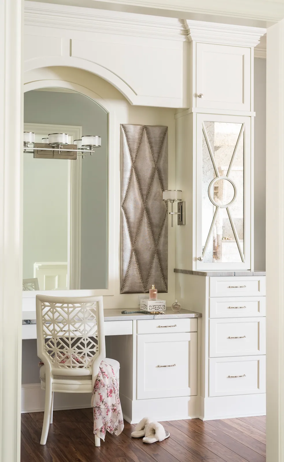 An elegant built-in white vanity area features an arched mirror, storage cabinets, and a decorative gray tufted panel. A patterned chair and slippers rest on the dark wood floor.