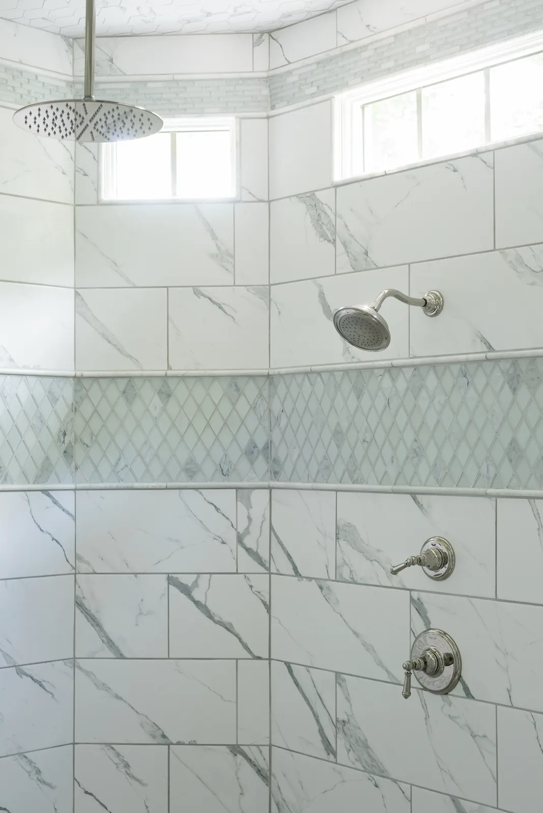 A modern, curved shower wall is covered in white marble-look tile, accented by a subtle gray diamond mosaic band and polished chrome shower fixtures.
