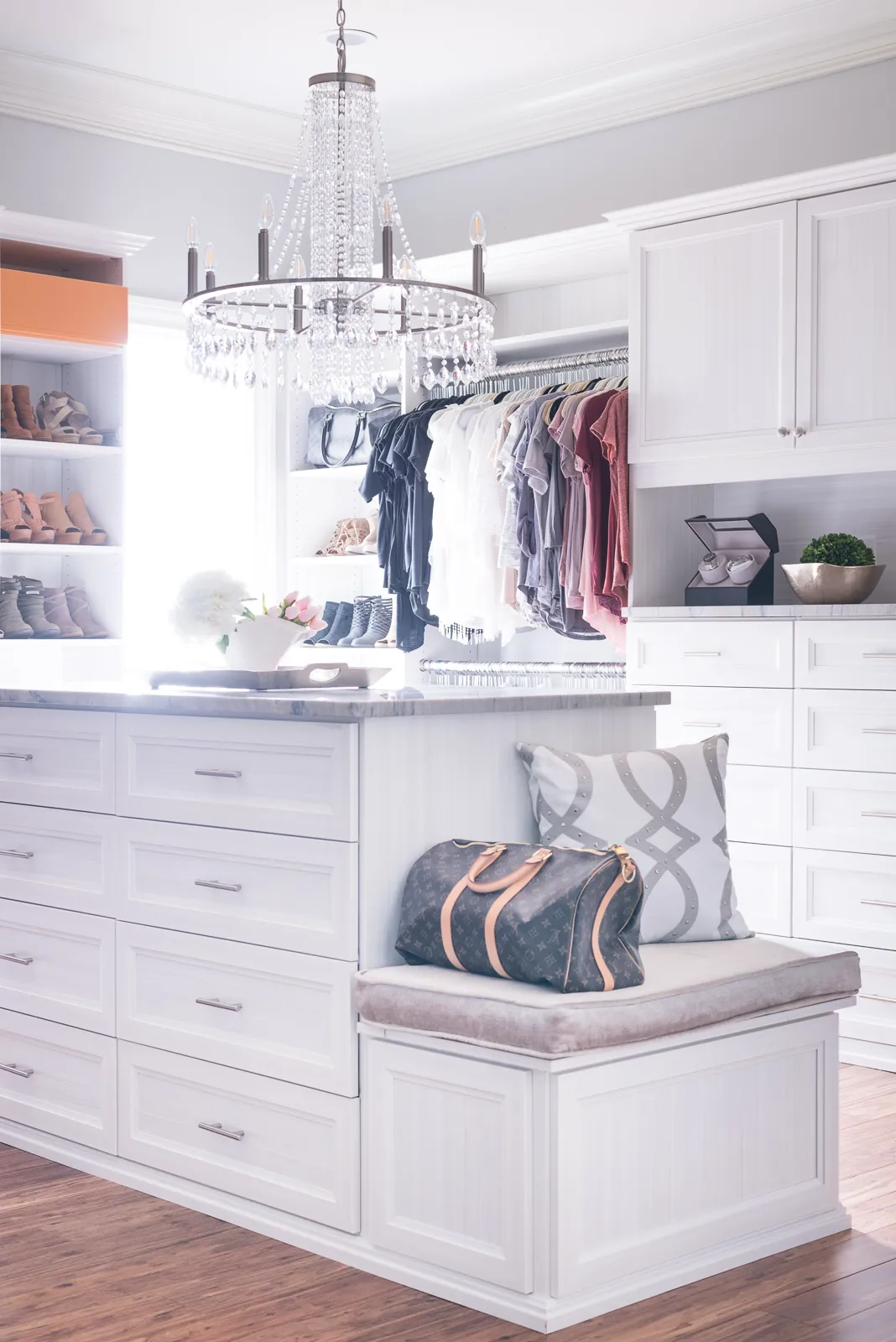 A bright, organized custom walk-in closet with white built-in storage, a central drawer island with a marble top, and a hanging crystal chandelier.