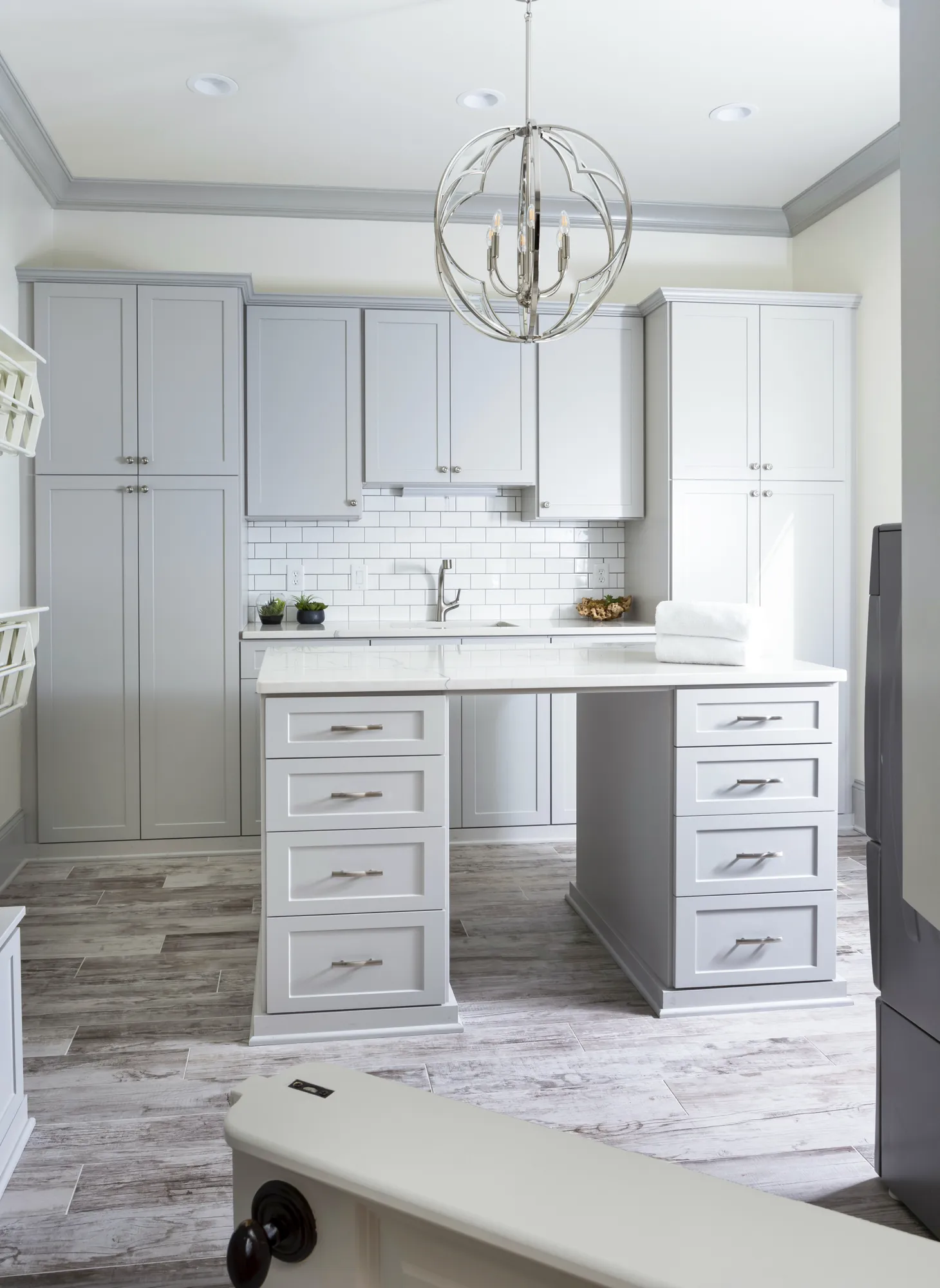 A luxury laundry room with light gray shaker cabinets, a subway tile backsplash, and a central white island counter for folding clothes.