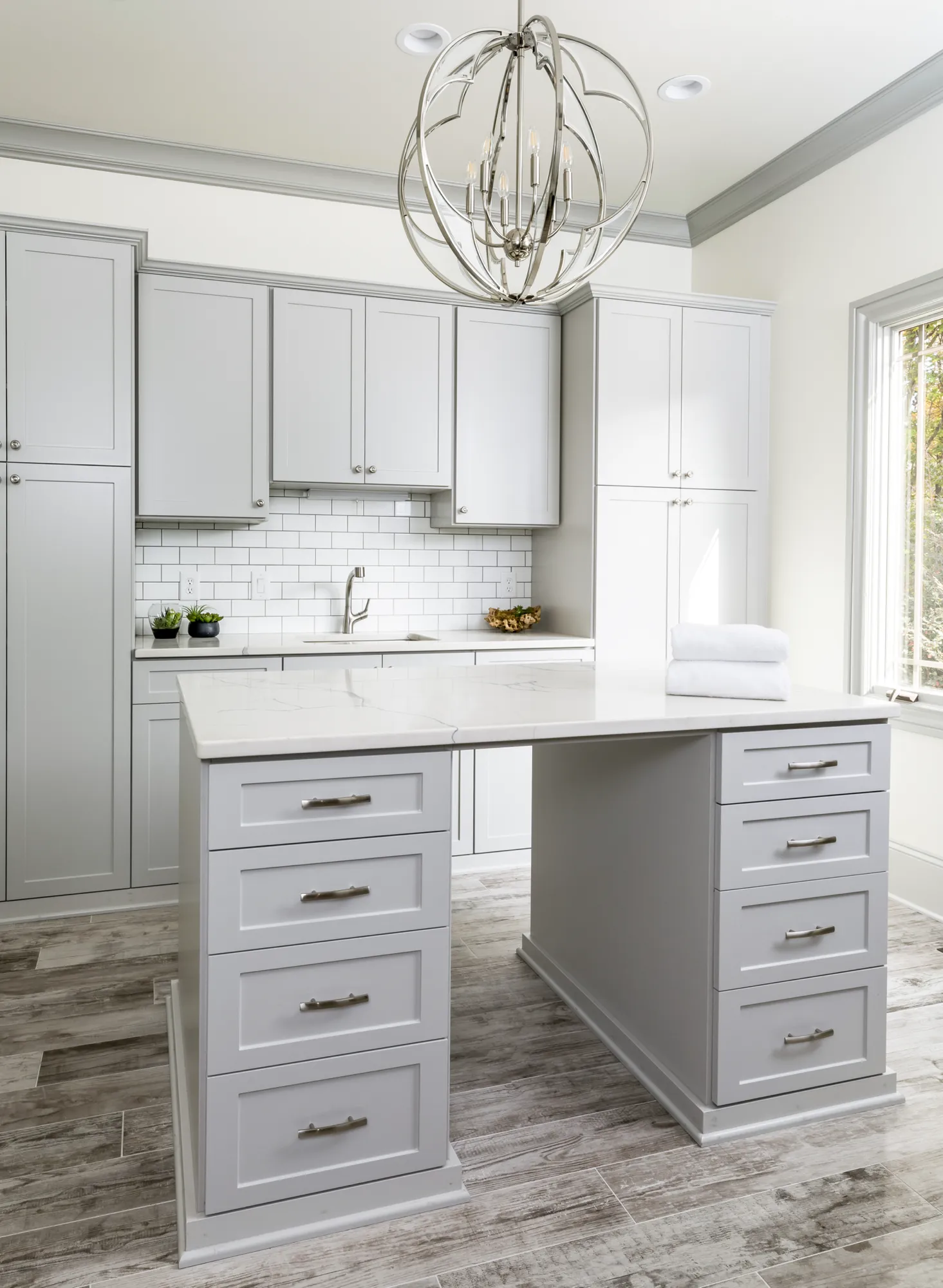A luxurious laundry room featuring light gray cabinetry, subway tile, and a large central island with a white marble countertop stacked white towels.
