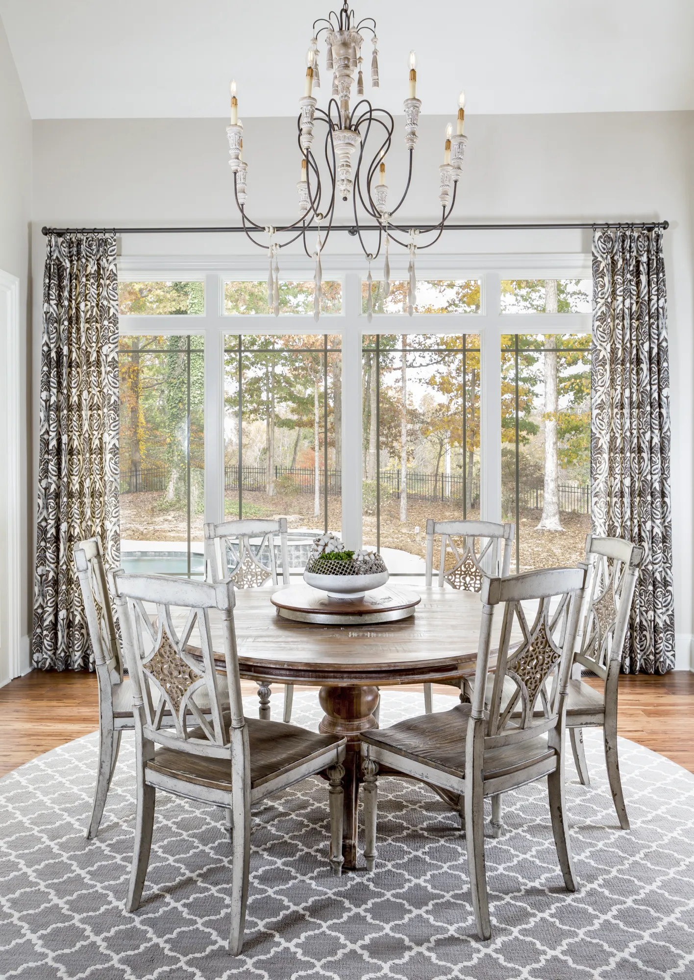 An elegant dining space featuring a round rustic wood table and distressed chairs on a grey patterned rug, framed by patterned curtains and a view of autumn trees.