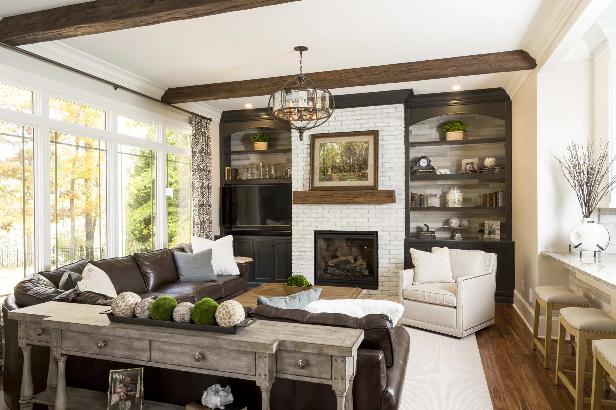 A stylish living room showcasing a white painted brick fireplace wall flanked by dark built-in shelves and topped with rustic ceiling beams.