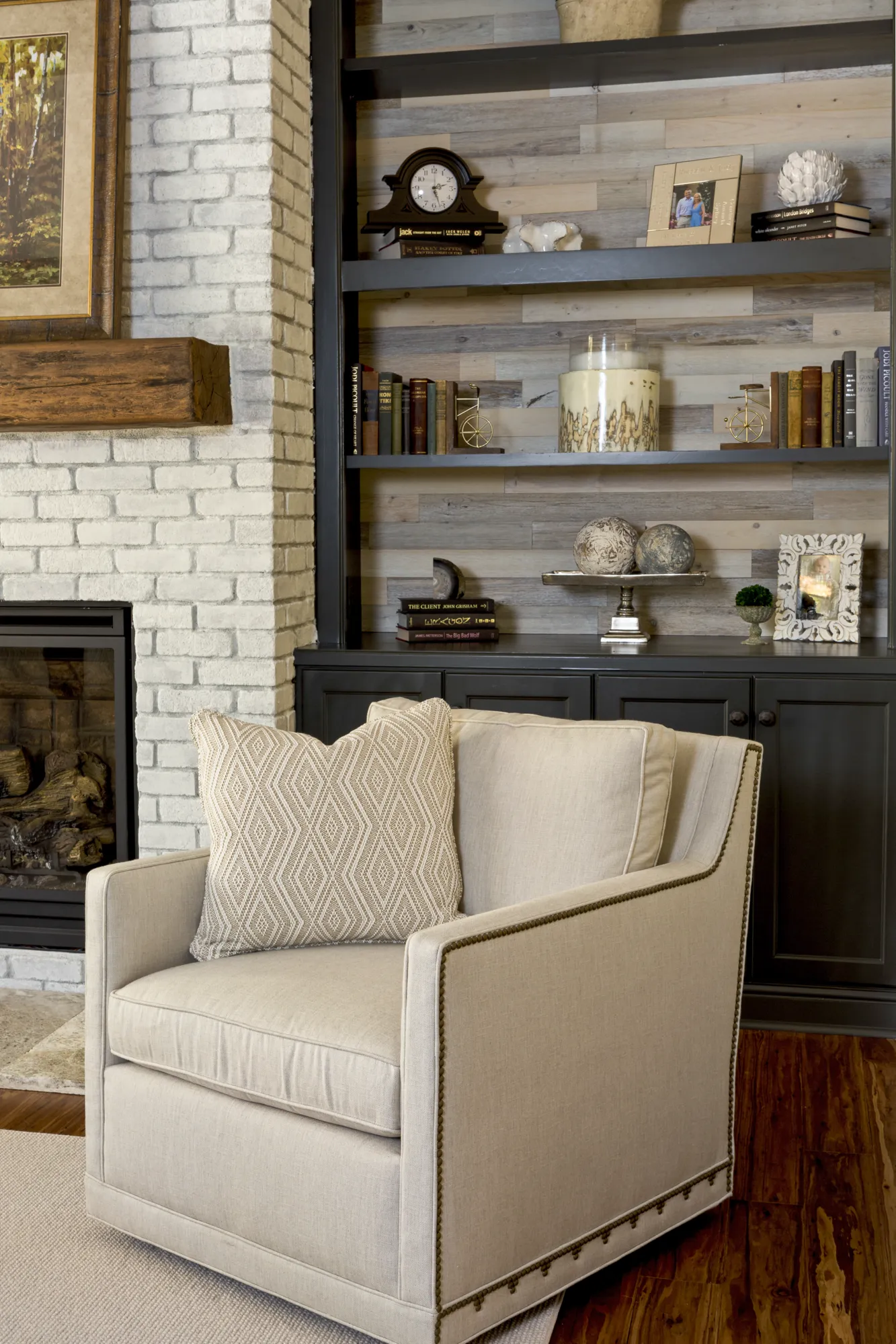 This cozy living room features a beige armchair next to a white brick fireplace and custom dark built-in shelving decorated with books and personal items.