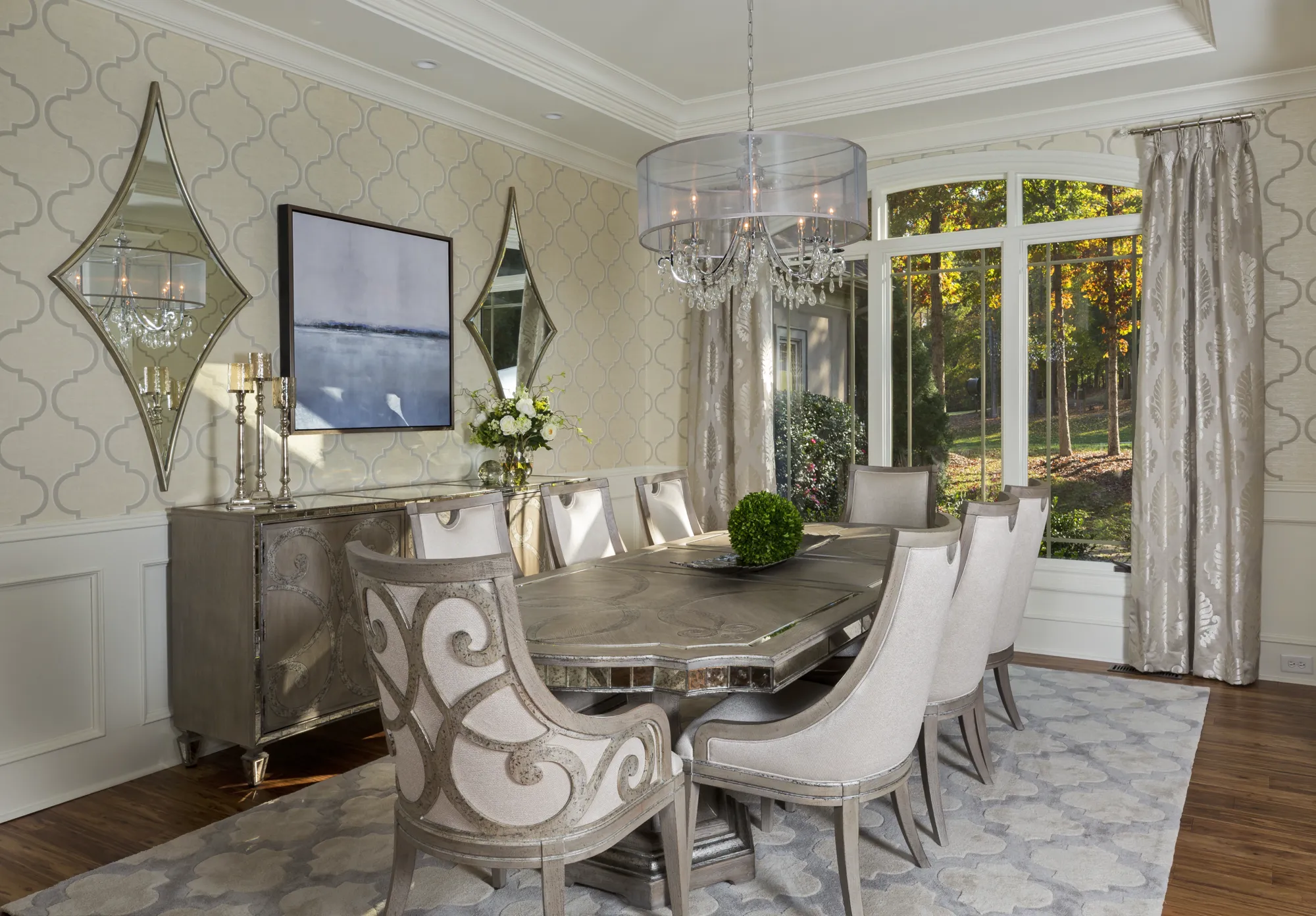 An elegant dining room featuring a metallic mirrored table and high-backed upholstered chairs. Diamond mirrors flank a console on patterned wallpaper, next to a large window overlooking a wooded area.
