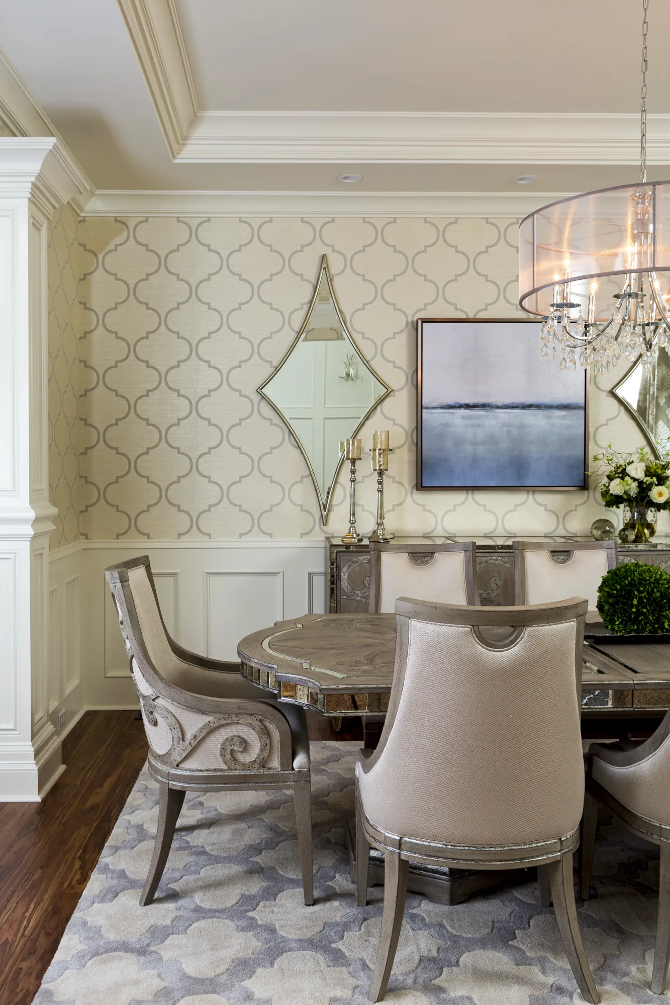 An elegant dining room featuring neutral upholstered chairs around a mirrored table. The patterned walls are accented by a landscape painting and mirrors above a buffet.