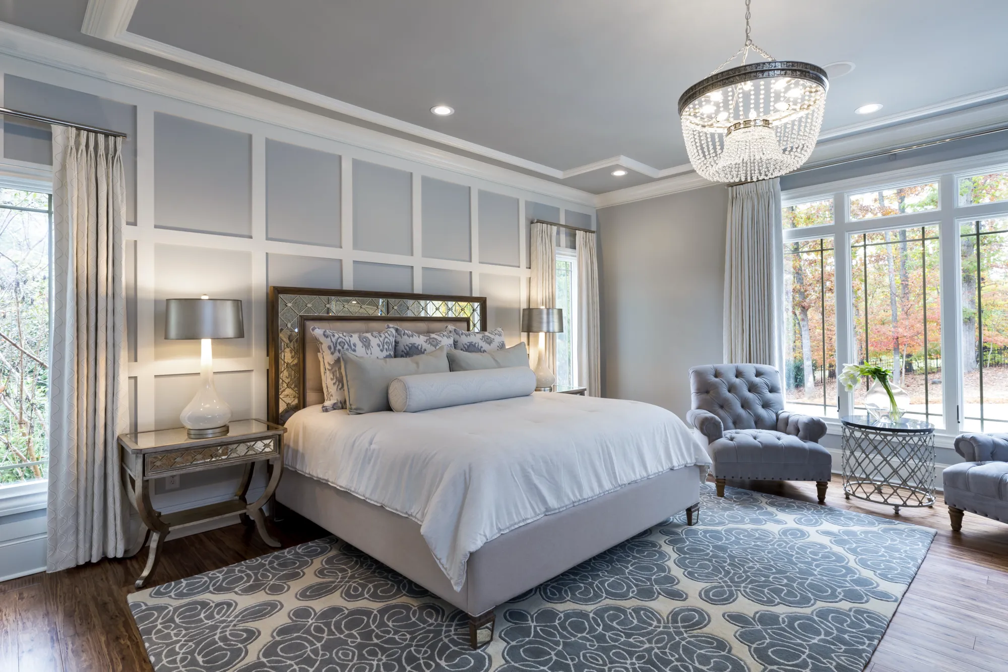 A beautifully designed bedroom featuring soft grey tones. The room has large windows, a bed with a mirrored headboard, a crystal chandelier, and a large grey patterned rug on hardwood floors.