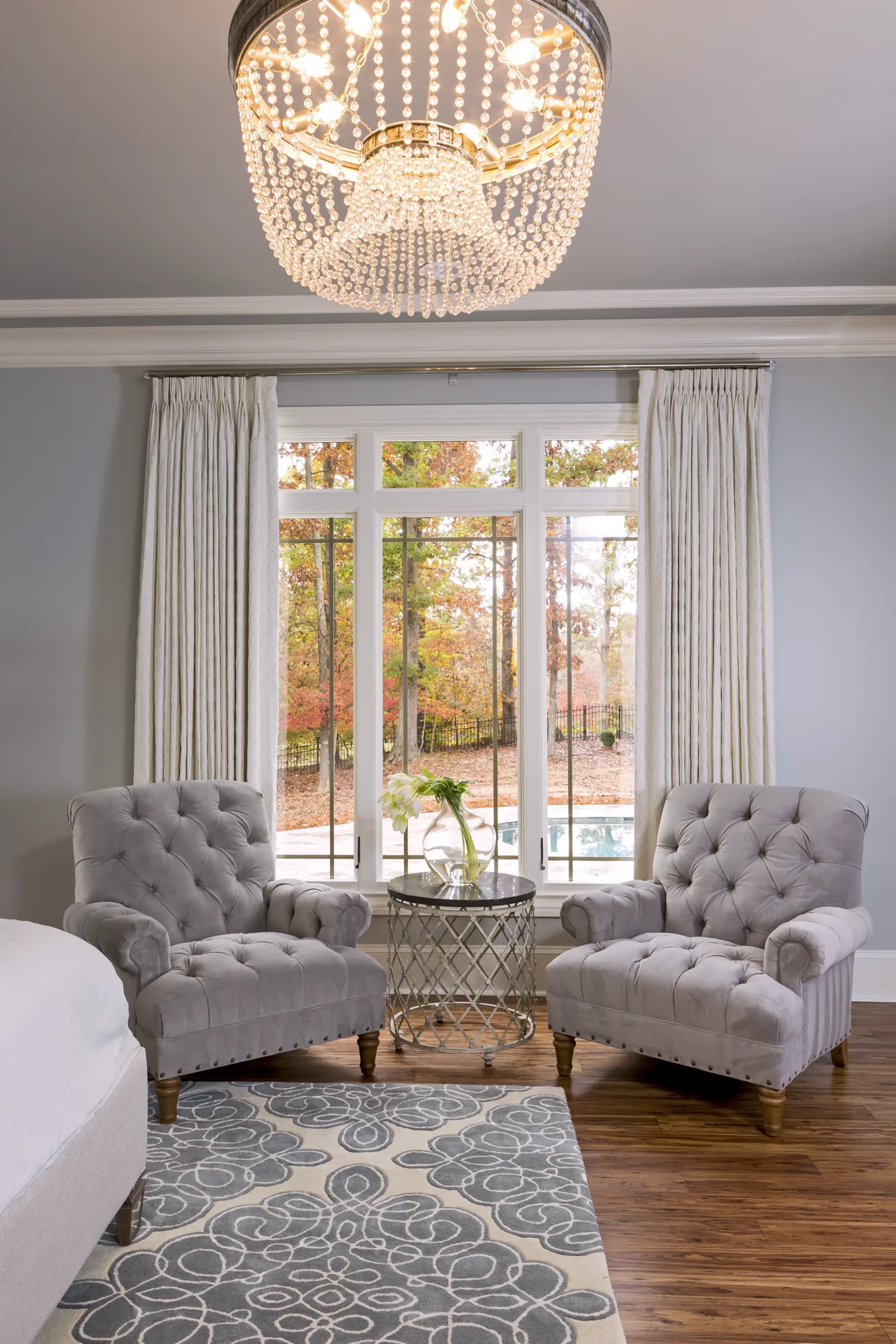 Two gray tufted velvet armchairs face a window with a view of autumn trees. A crystal chandelier hangs above a patterned rug and glass side table.