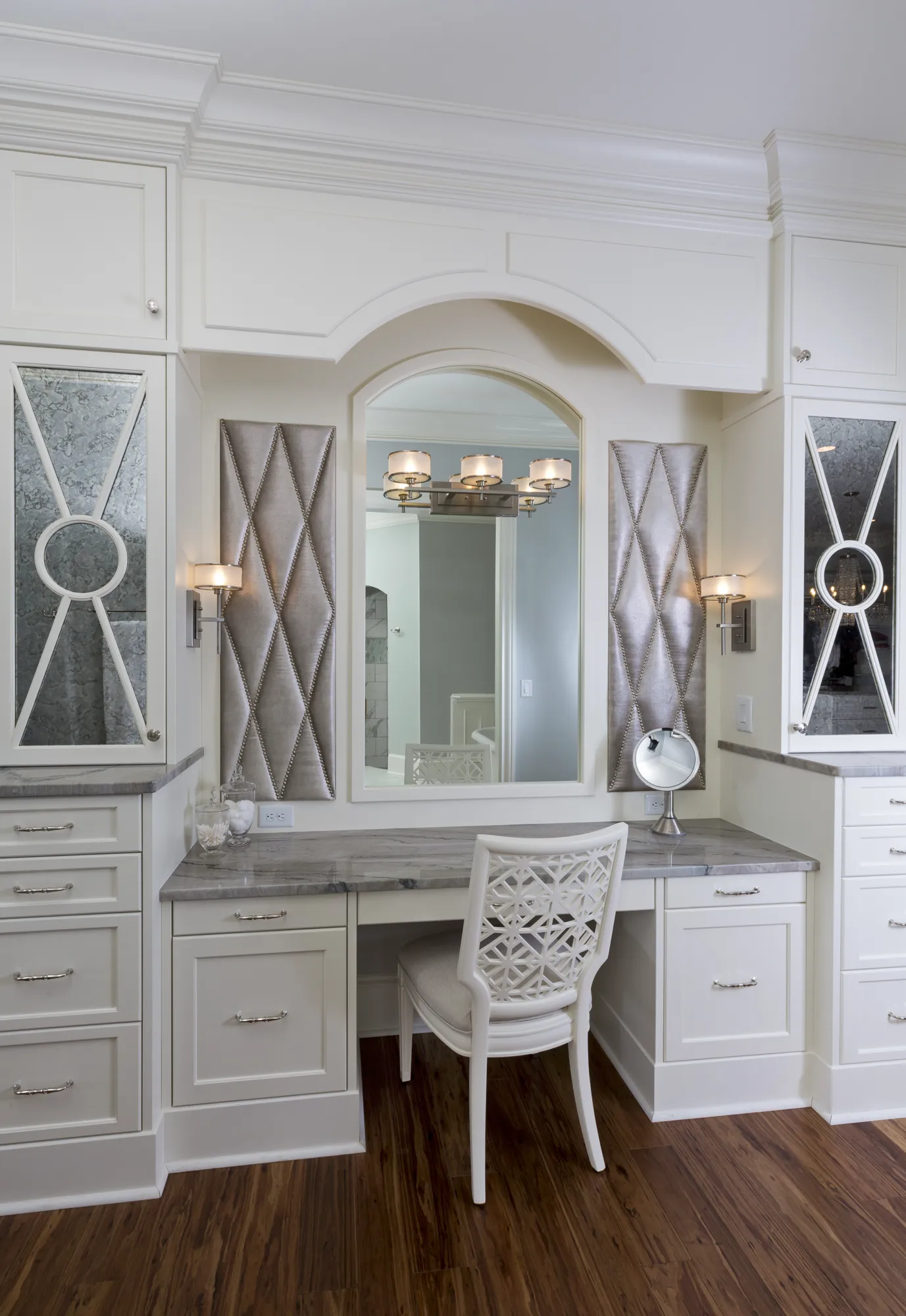 A bright, elegant custom built-in vanity station with white cabinetry, a grey marble counter, and dark wood floors. It features an arched mirror flanked by diamond-quilted wall panels and sconces.