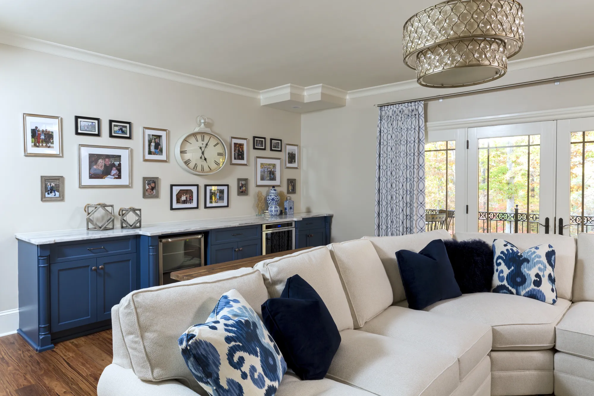 An elegant living room featuring a cream sectional sofa with blue pillows in front of a navy built-in cabinet and photo gallery wall. French doors are on the right.