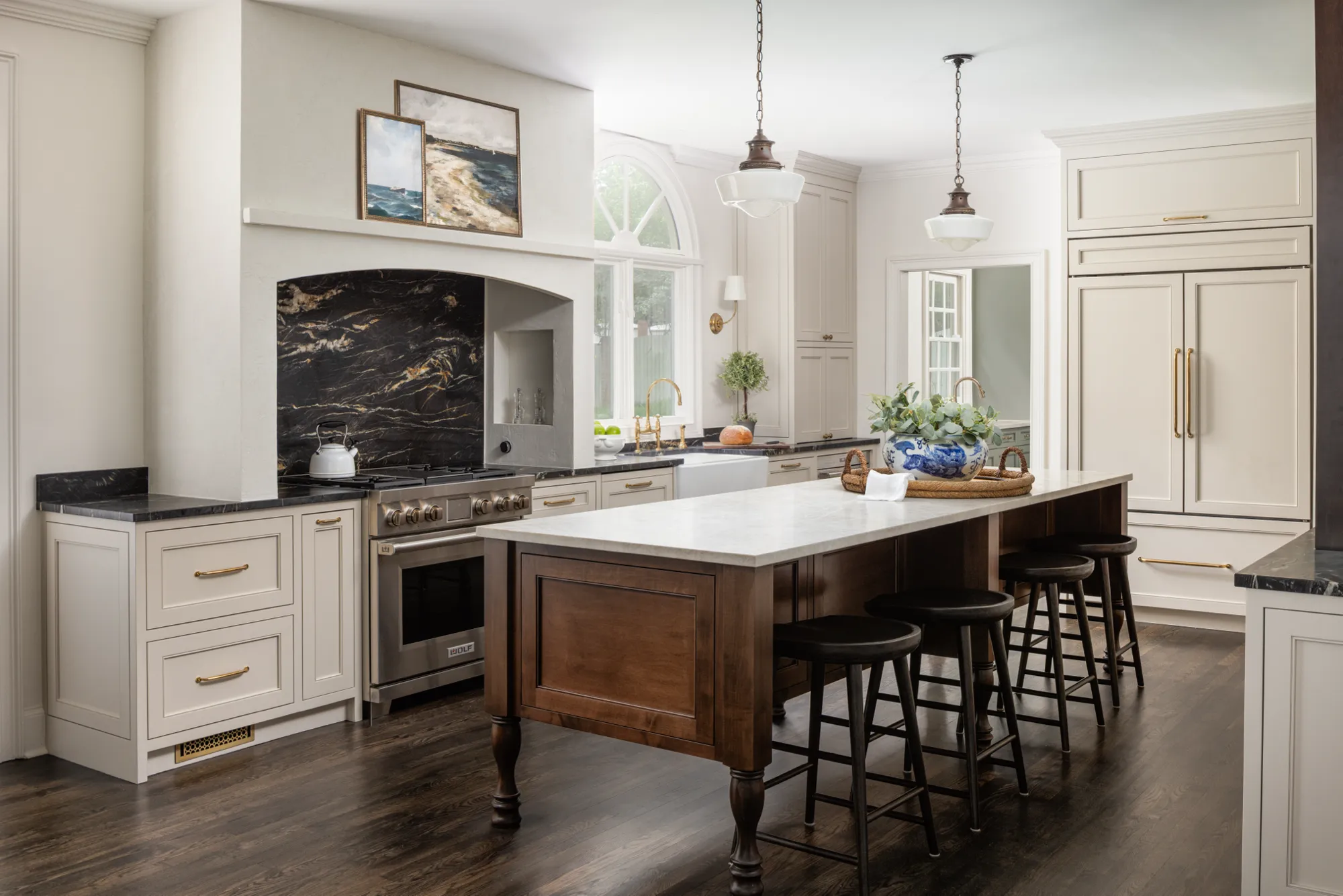 A bright, elegant kitchen with cream cabinets, dark wood floors, and a large central island with four stools. Black marbled stone frames the professional range.