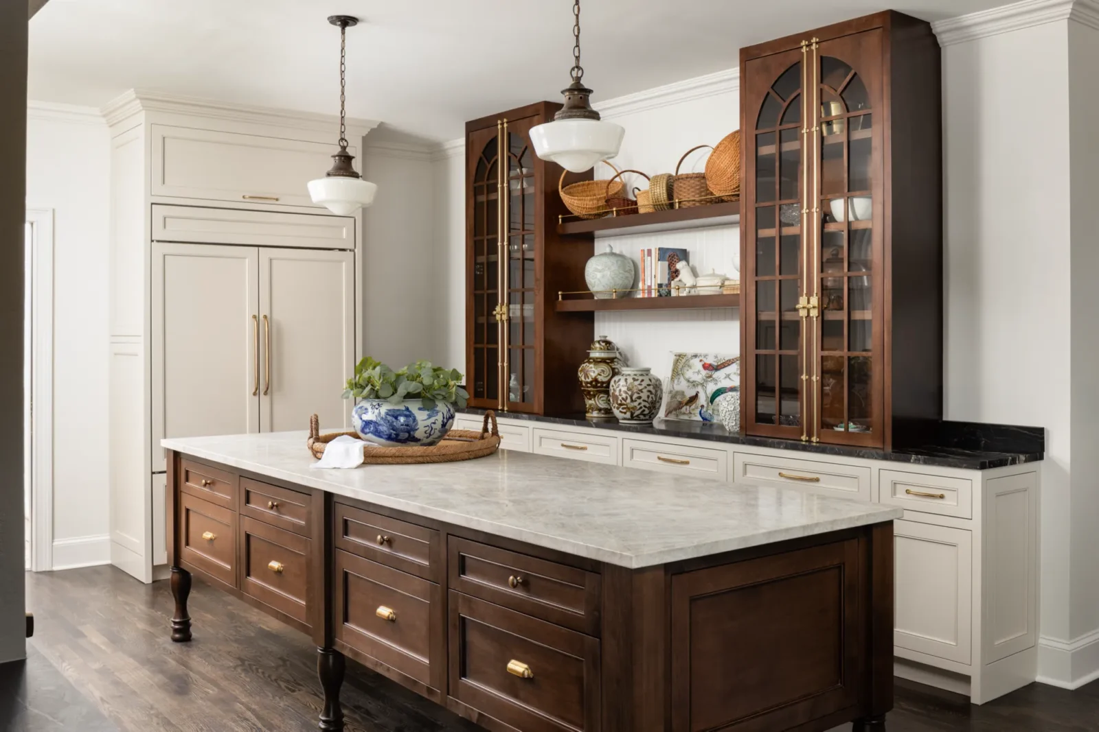 An elegant kitchen features a dark wood island with a light marble countertop. Cream and dark wood cabinetry surrounds open shelves decorated with pottery, baskets, and greenery.