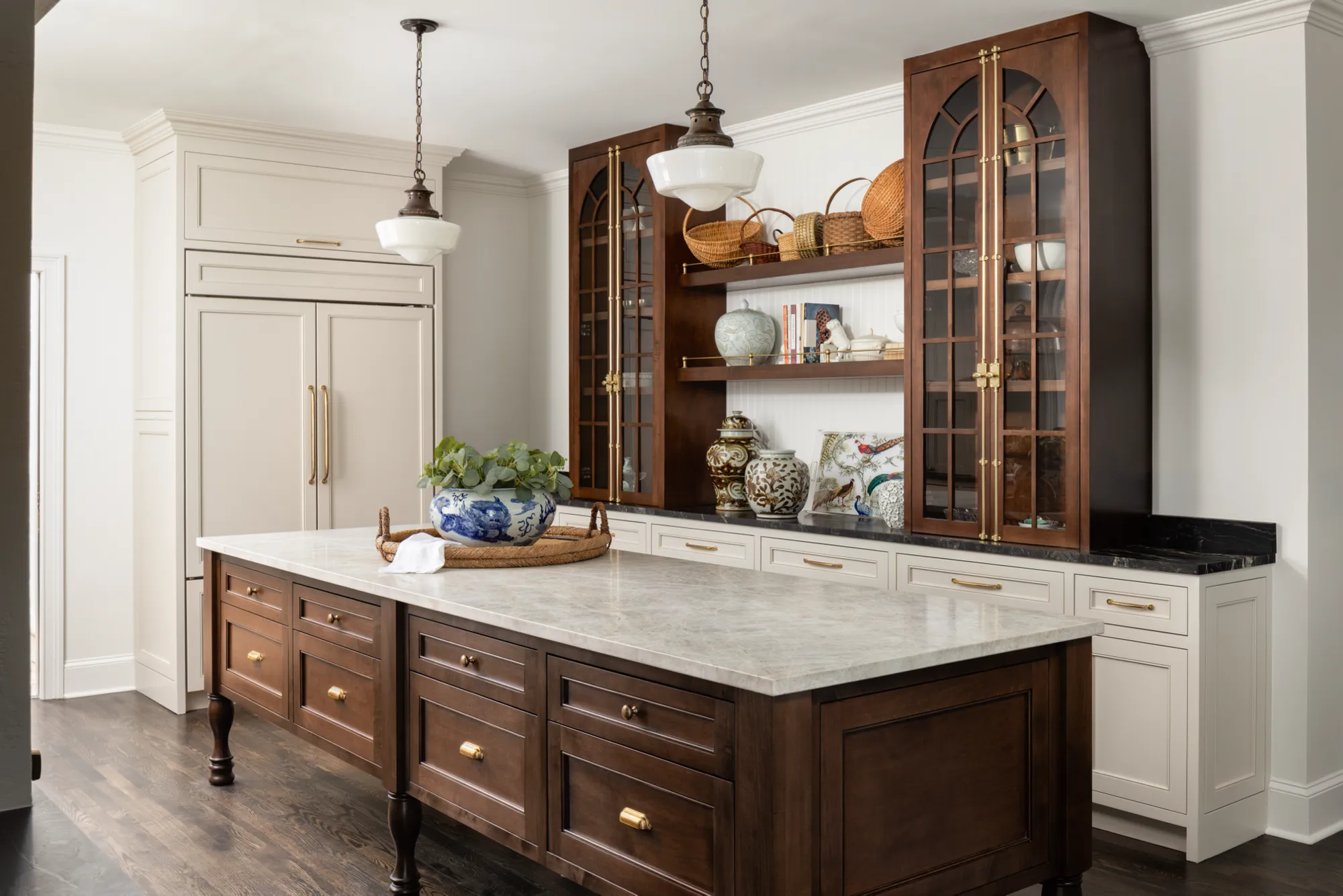 An elegant kitchen features a dark wood island with a light marble countertop. Cream and dark wood cabinetry surrounds open shelves decorated with pottery, baskets, and greenery.