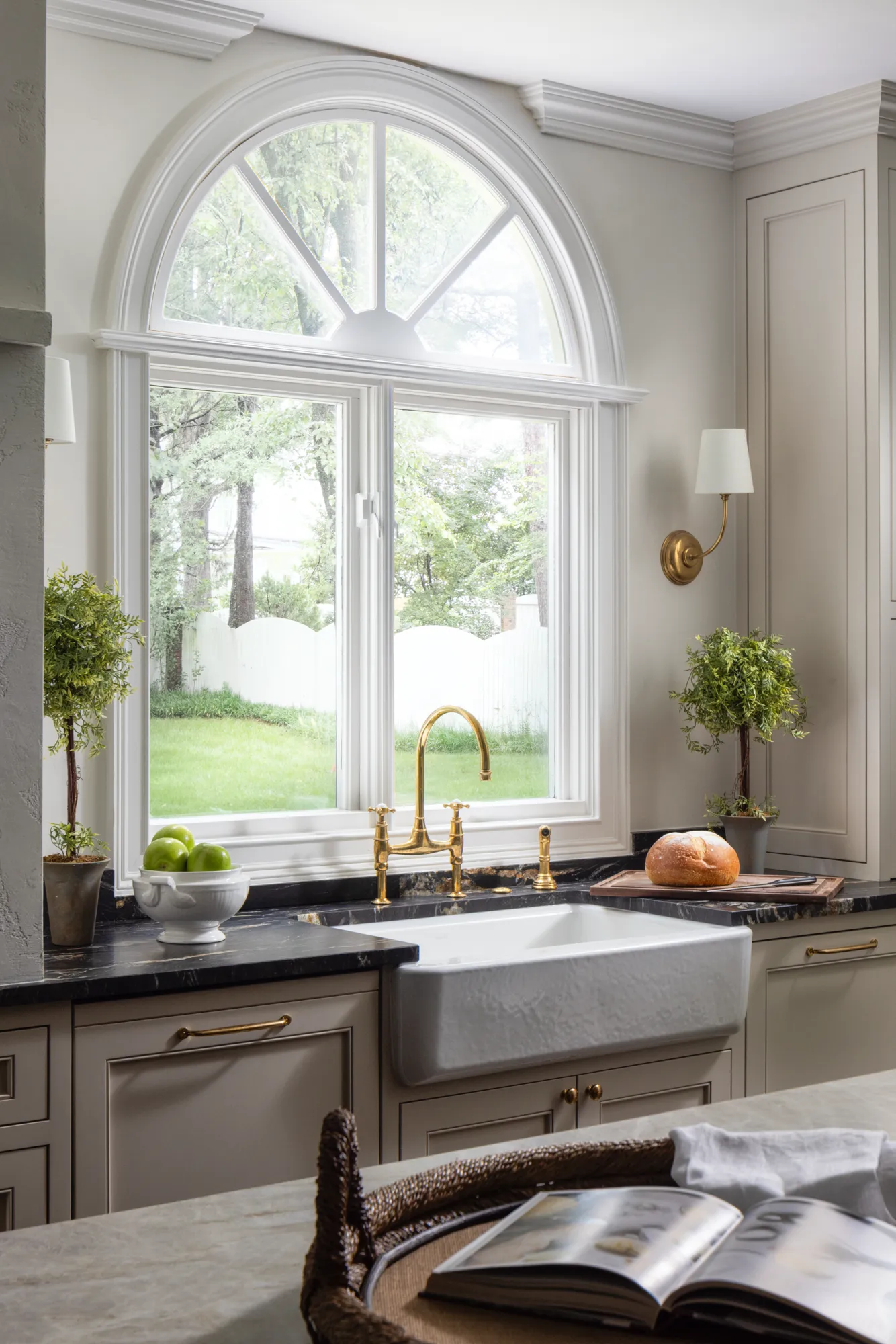 A kitchen featuring a large white farmhouse sink, brass faucet, and dark countertop beneath a bright, arched window overlooking a green yard.