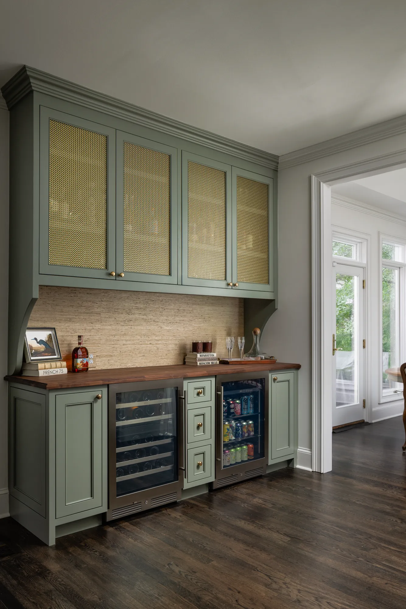 A built-in bar area featuring sage green custom cabinetry with brass mesh inserts and a dark wood countertop. It includes dual under-counter wine and beverage refrigerators.