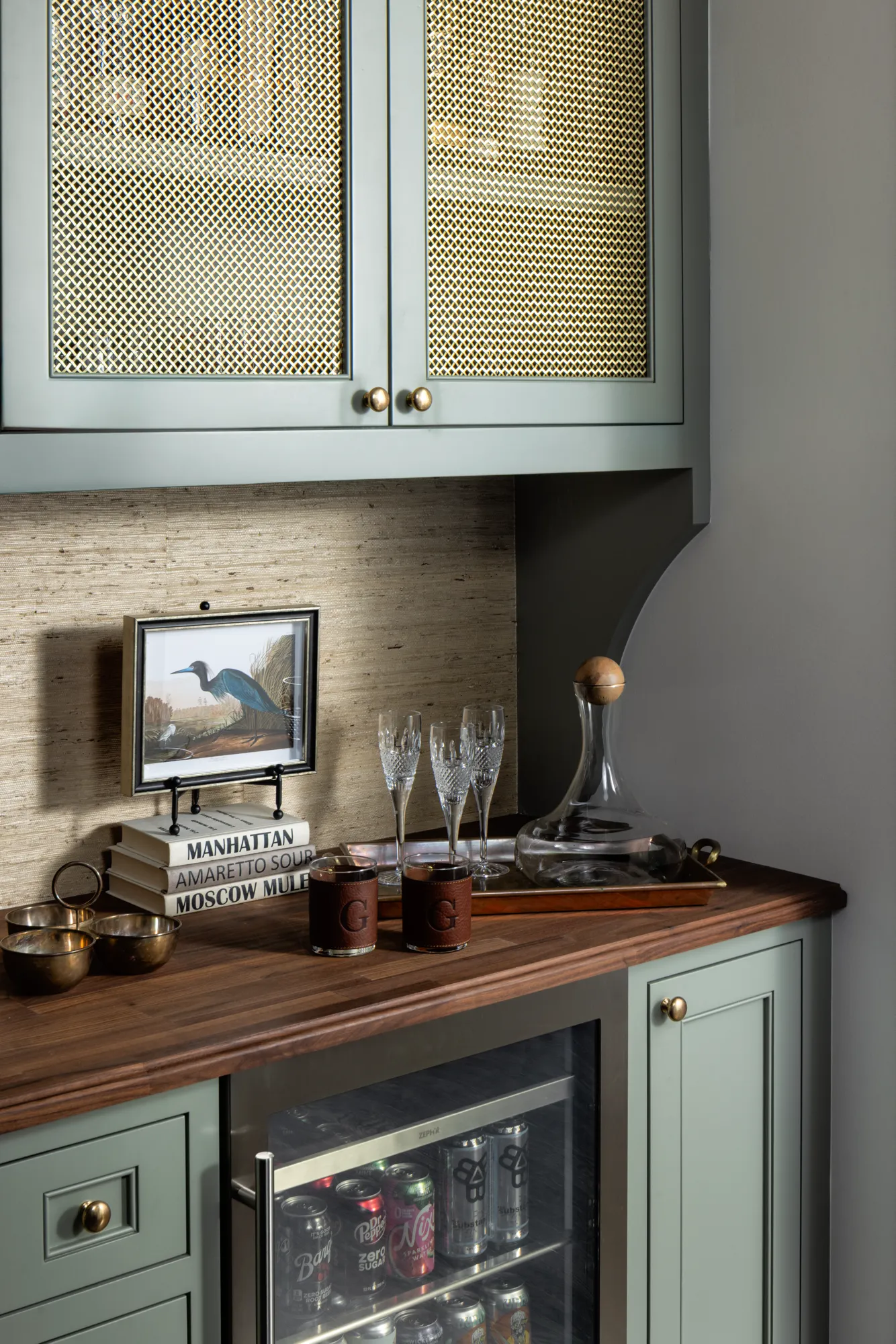 A sophisticated built-in bar area featuring sage green cabinets with woven lattice inserts and a wood counter, styled with glassware, a decanter, and cocktail books.