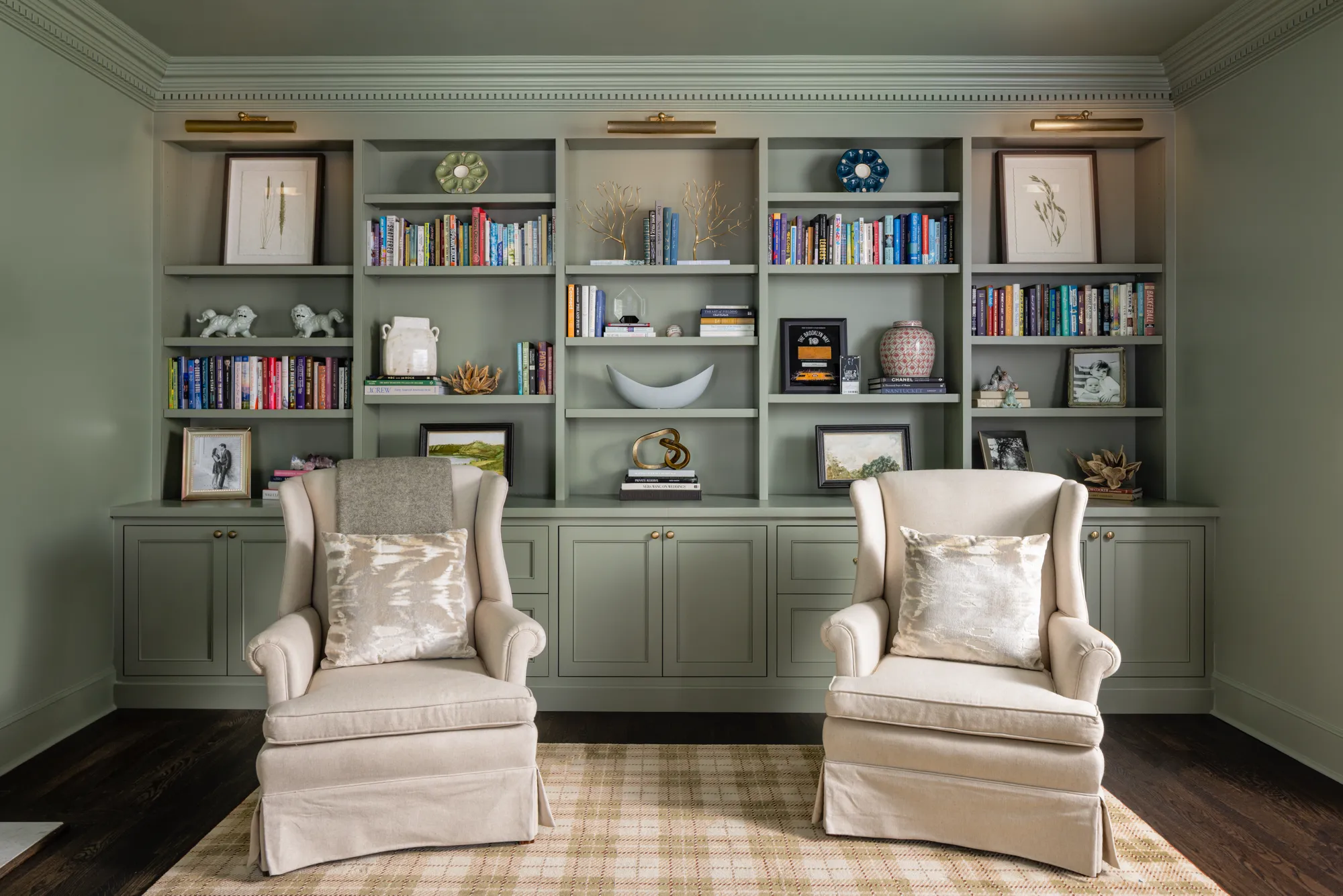Two cream wingback armchairs are centered on a plaid rug before floor-to-ceiling sage green built-in bookshelves filled with books and decorative art.