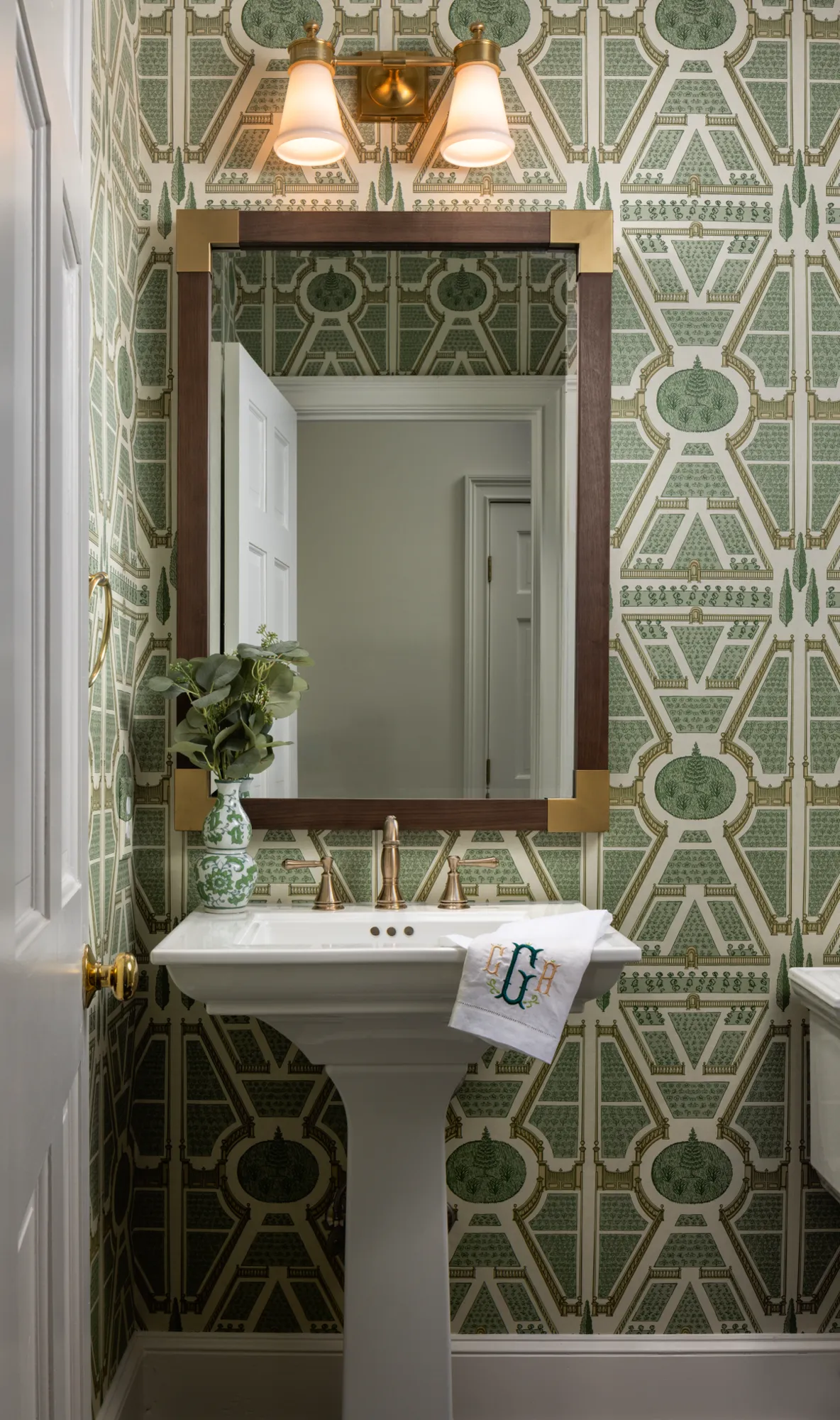 A bathroom with green patterned garden wallpaper, a white pedestal sink, and brass fixtures. A wood-framed mirror hangs above the sink.