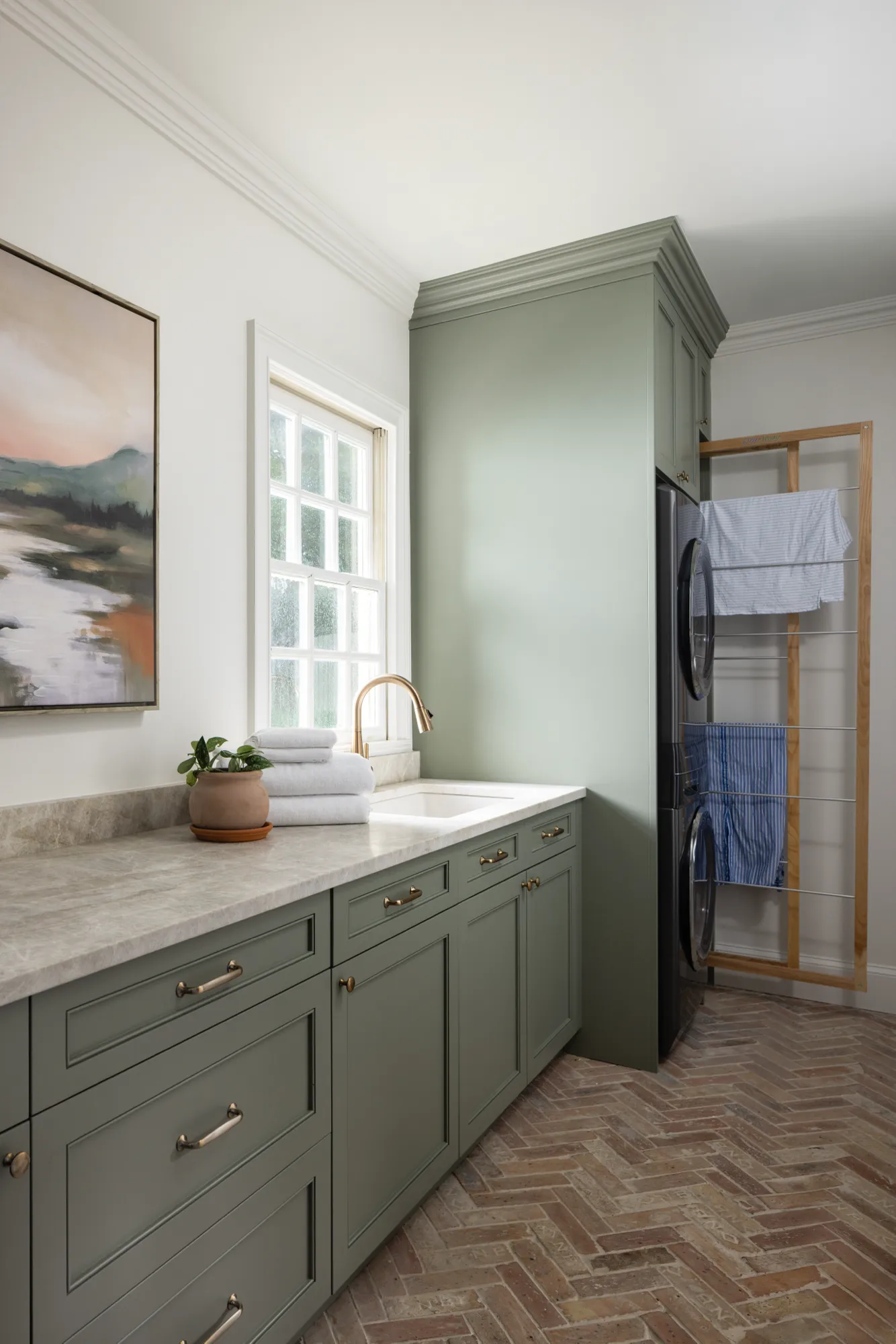 A stylish laundry room featuring sage green cabinetry, a marble countertop, and a brass faucet. A stacked washer and dryer sit next to a wooden drying rack, set above brick herringbone flooring.