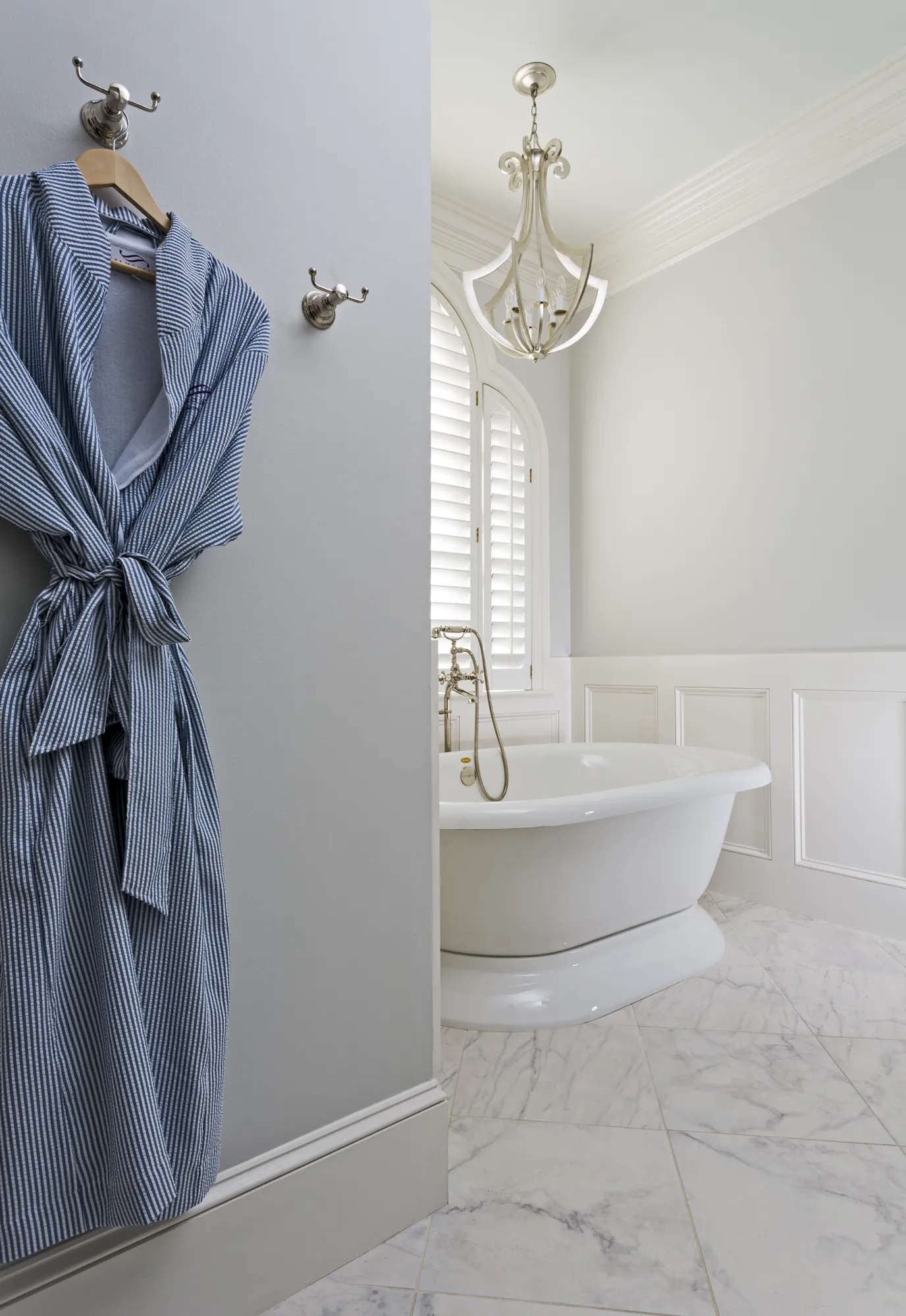 A blue and white striped robe hangs on a wall, partially revealing a luxurious bathroom with a white freestanding tub and marble floor.