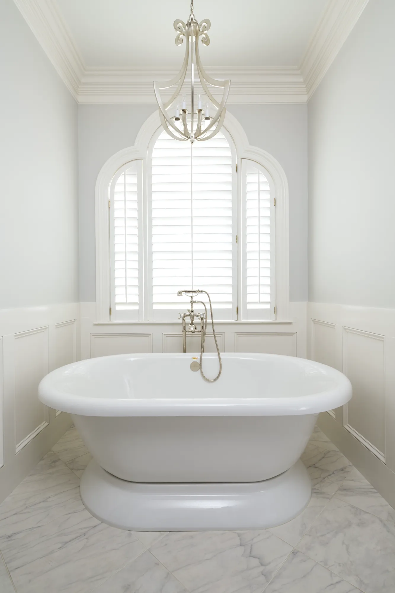 A luxurious white bathroom with a pedestal soaking tub and chrome fixtures centered under an arched window with shutters. The room features marble tile floors and an ornate chandelier.