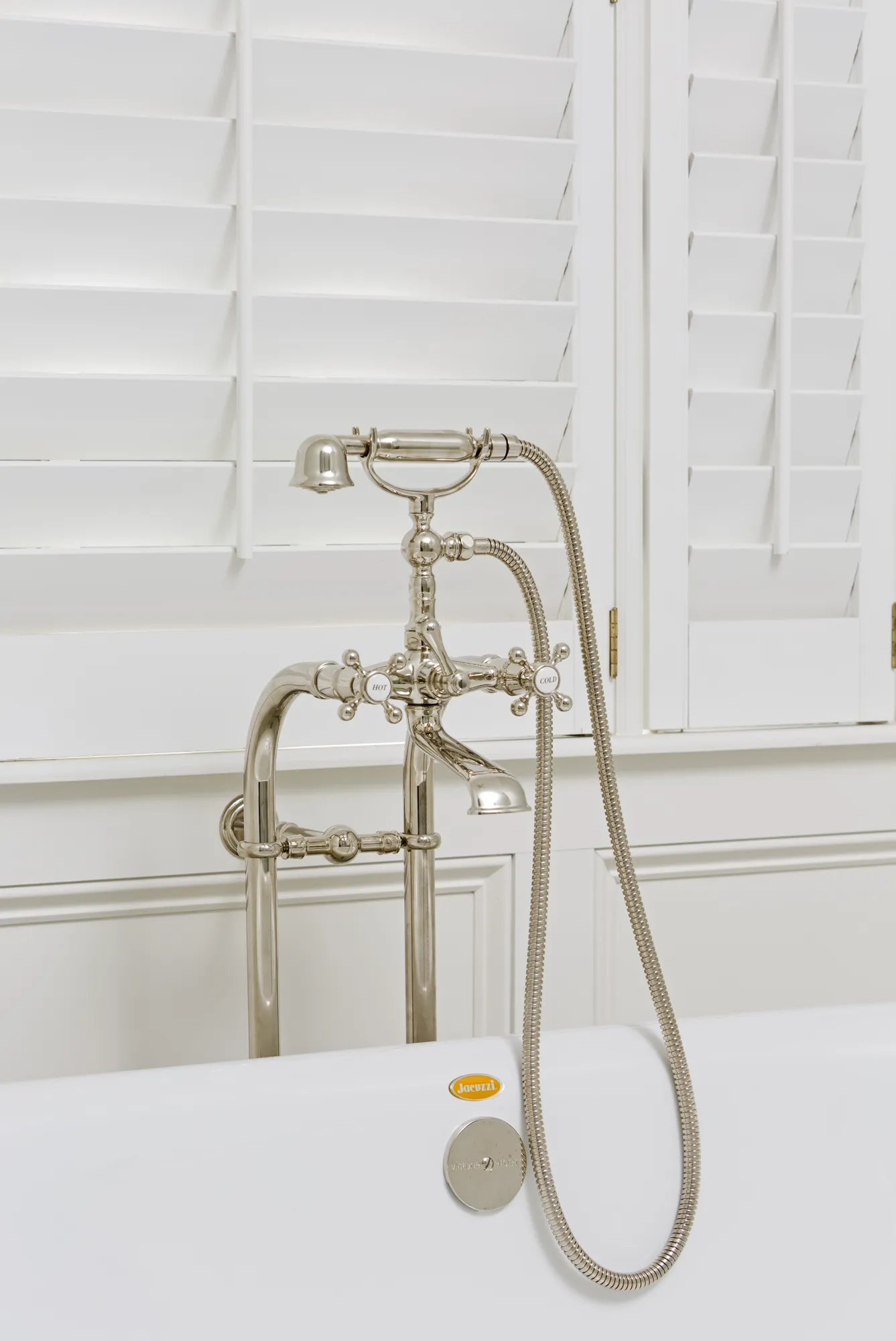 A polished metal freestanding bath faucet with cross-handles and a handheld shower stands in a clean white bathroom with plantation shutters.