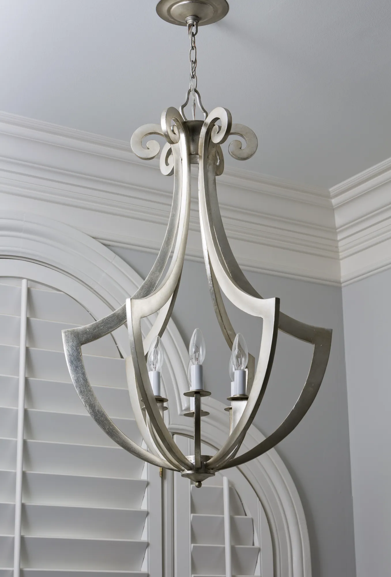 A modern silver chandelier featuring a scrollwork cage design and exposed candle bulbs hangs above arched windows with white plantation shutters.