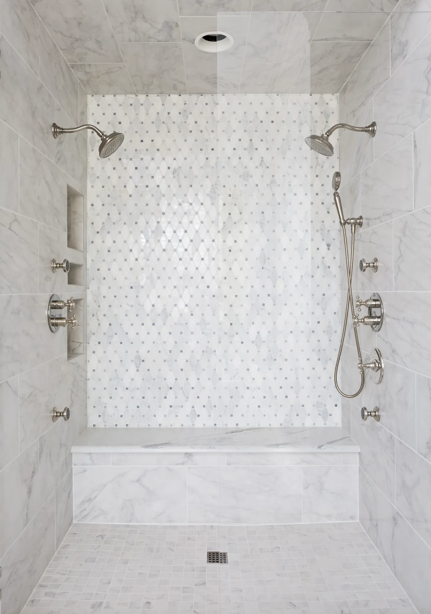 An elegant walk-in shower with white marble tiling, featuring a central diamond mosaic accent wall, a built-in bench, and polished chrome fixtures.