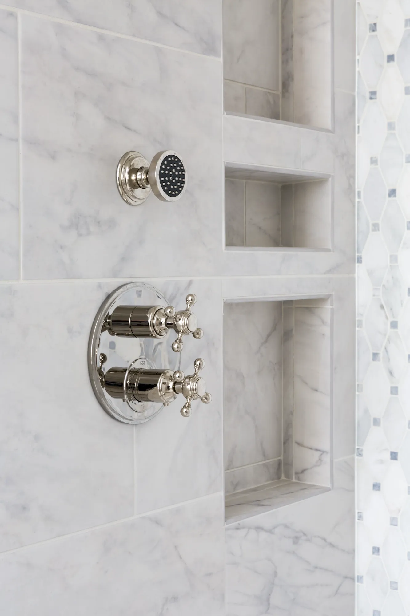 Polished nickel shower fixtures, including a small round head and cross-handle controls, are mounted on a white marble tile wall with three built-in storage niches.
